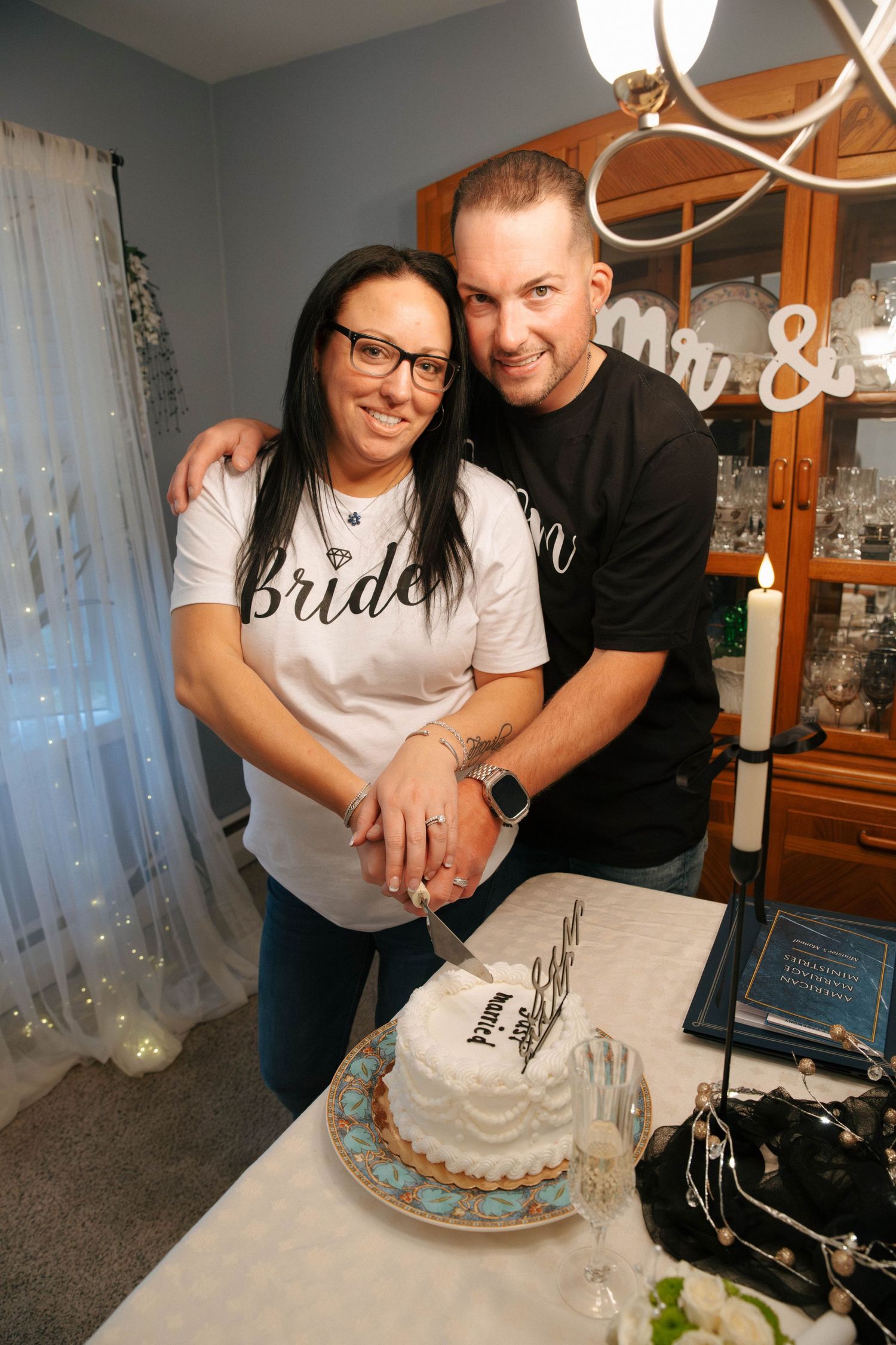 Couple in Bride and Groom shirts cutting a wedding cake together at a celebration party.