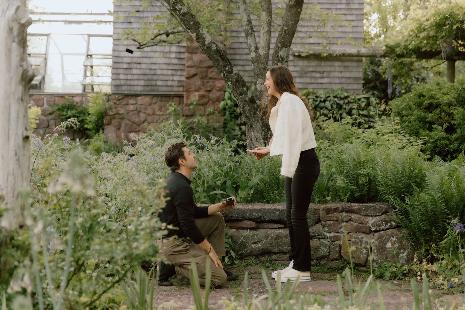 Man on one knee proposing to his partner in a beautiful garden setting surrounded by greenery.