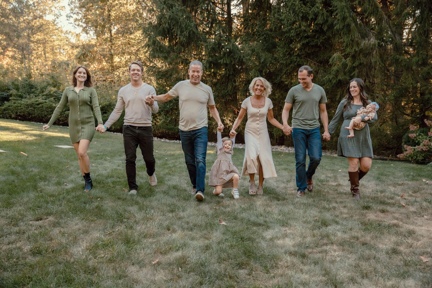 Happy extended family walking together across green lawn in front of tall evergreen trees.