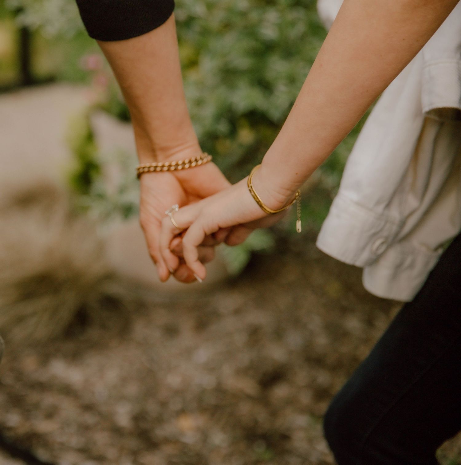 Two people holding hands outdoors, both wearing gold bracelets, surrounded by lush greenery.