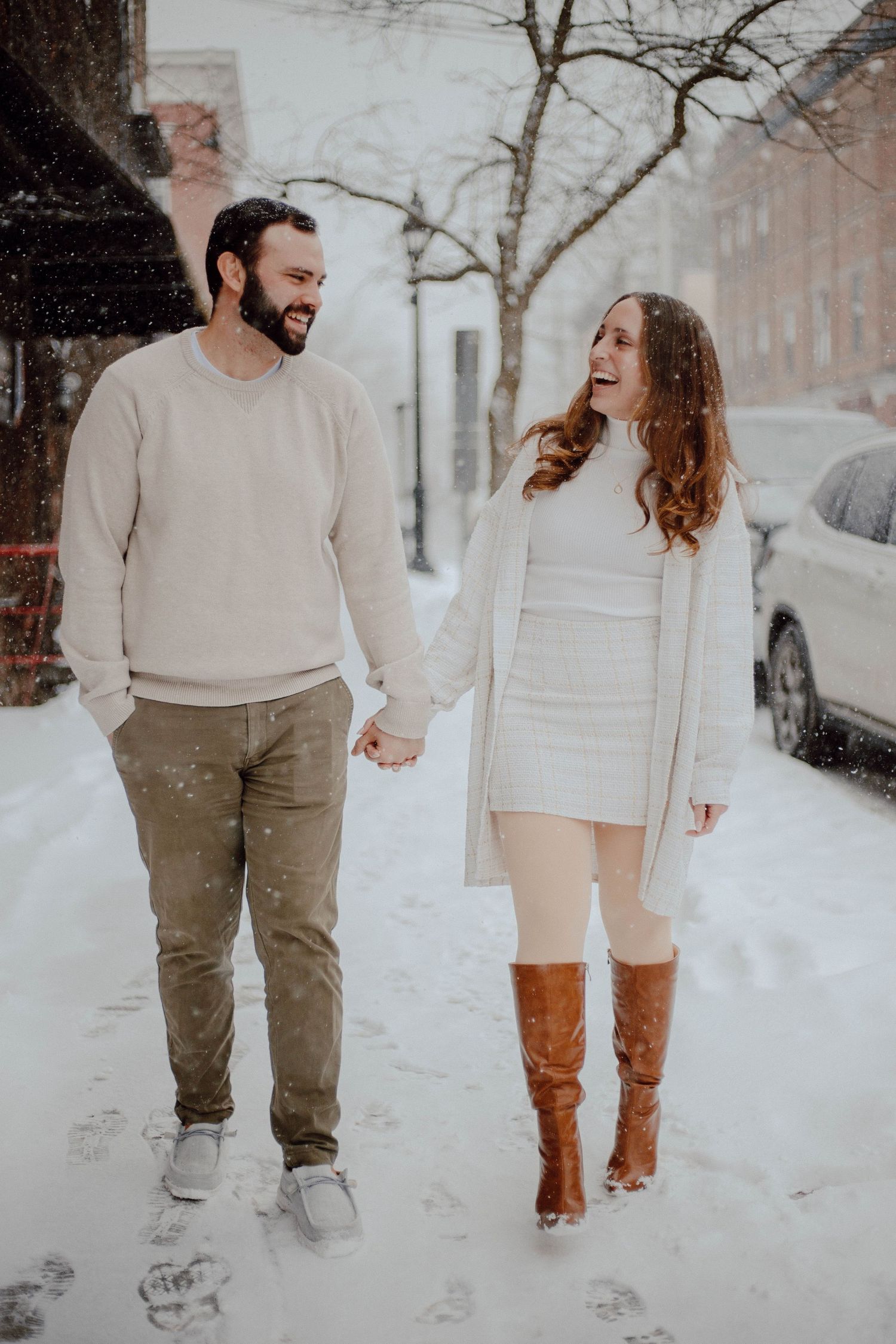 Couple holding hands and laughing while walking on a snowy city street in winter outfits.