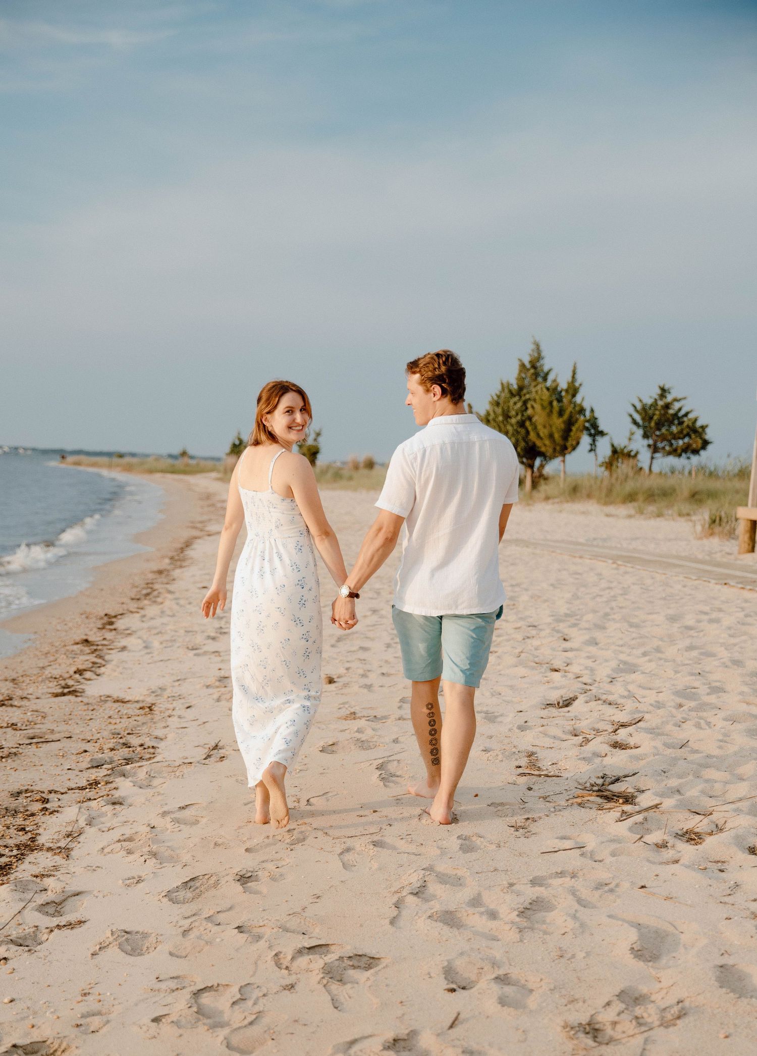 Happy couple walking hand in hand along a sandy beach, woman looking back and smiling.