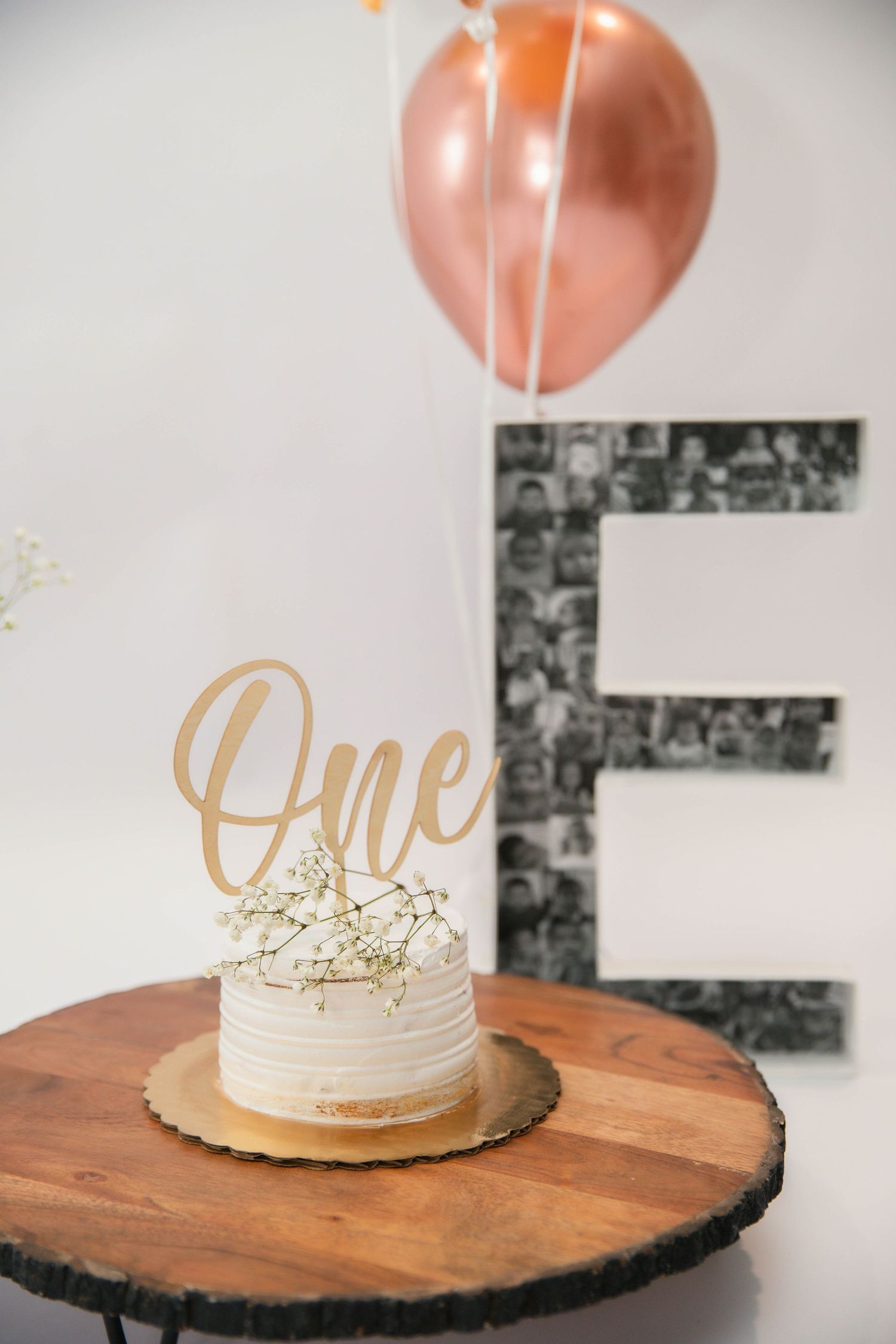 White frosted first birthday smash cake with gold 'One' topper on wood slice, rose gold balloon and letter E backdrop.