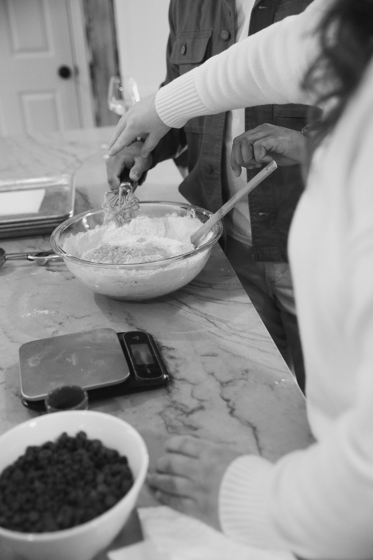 Two people mixing batter in a glass bowl on a marble counter with a kitchen scale and berries nearby.
