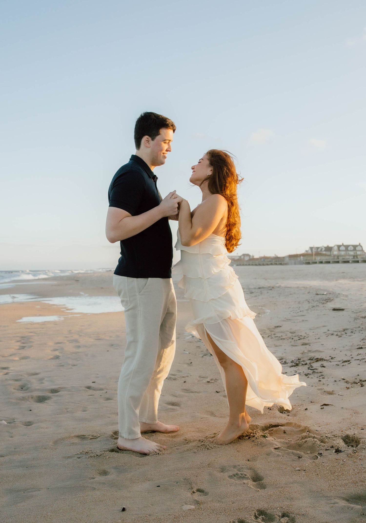 Couple dancing barefoot on a sandy beach at sunset, woman in white dress, man in black shirt and khaki pants.