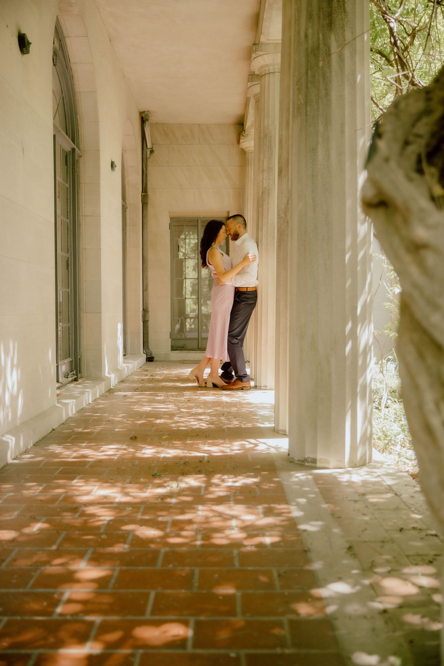 Couple sharing a romantic embrace in a sunlit colonial-style covered porch with brick flooring.
