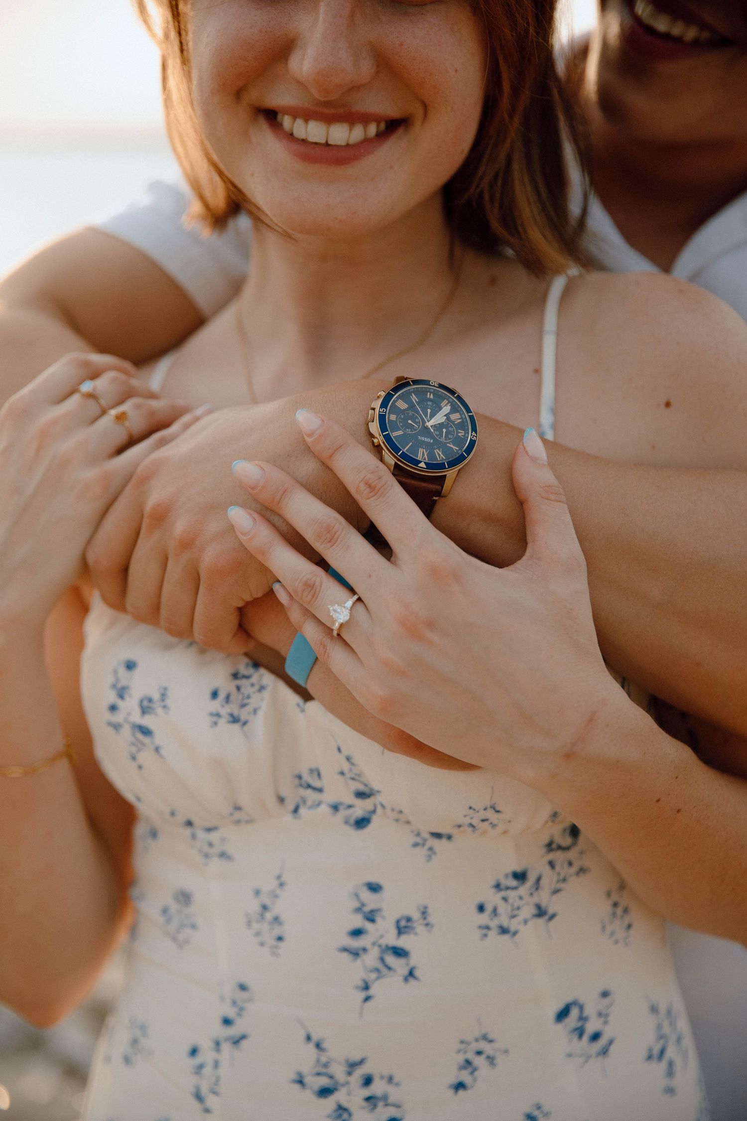 Smiling woman in floral dress with engagement ring, embraced by partner wearing a blue chronograph watch.