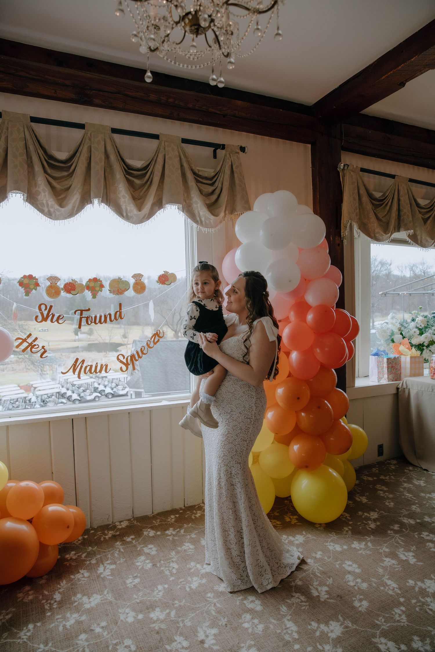 Bride in lace gown holding toddler near colorful balloon arch with She Found Her Main Sauce sign.