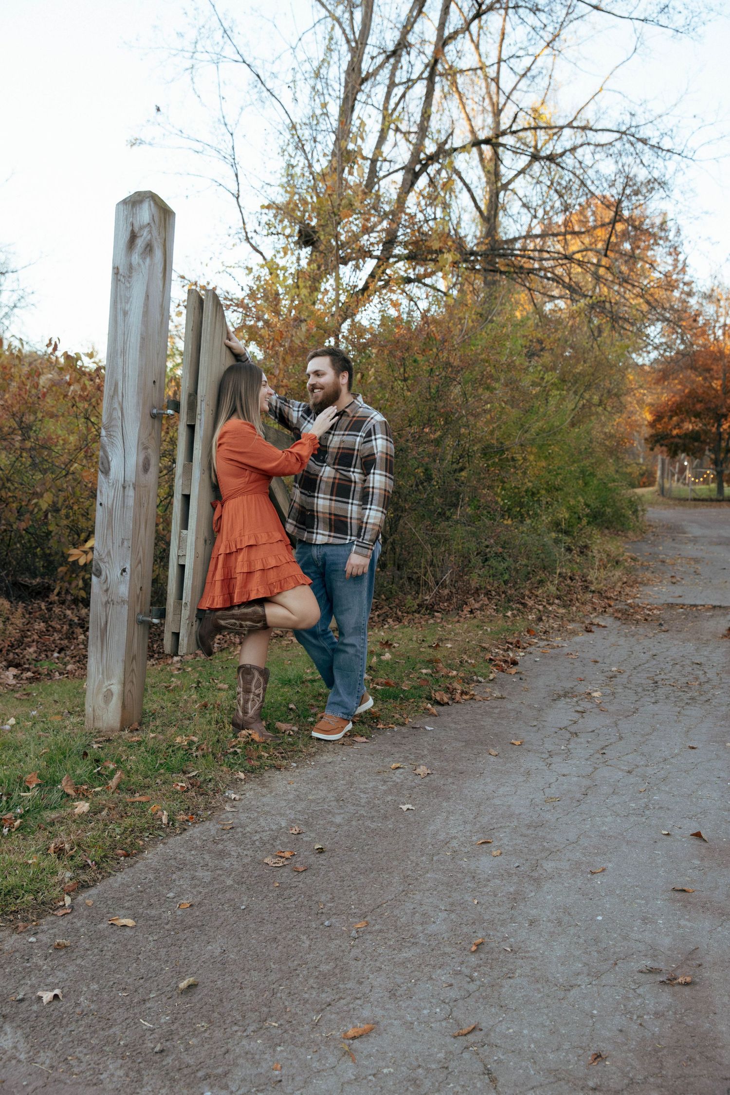 Couple sharing a romantic moment by a wooden gate on a scenic autumn path, woman in orange dress, man in plaid shirt.