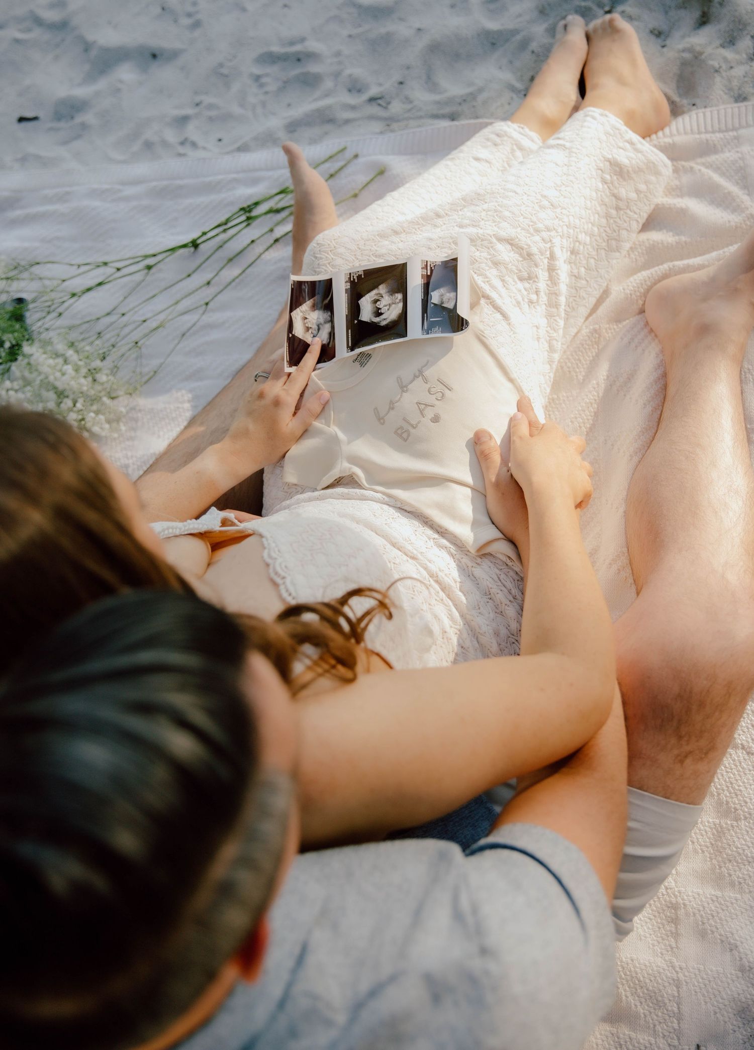 Couple lying on sand looking at ultrasound photos during a beach maternity announcement session.