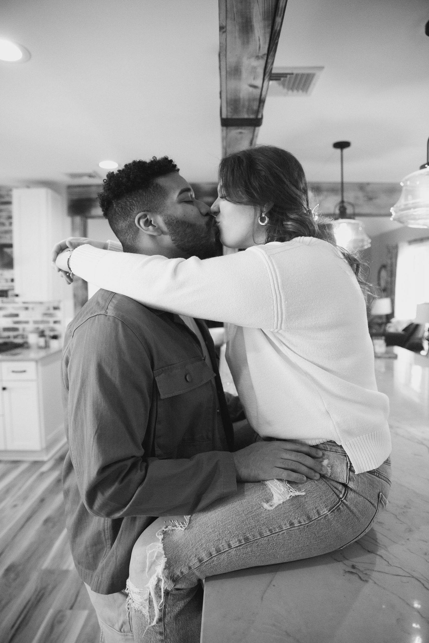Black and white photo of an interracial couple sharing a romantic kiss in a modern kitchen interior.