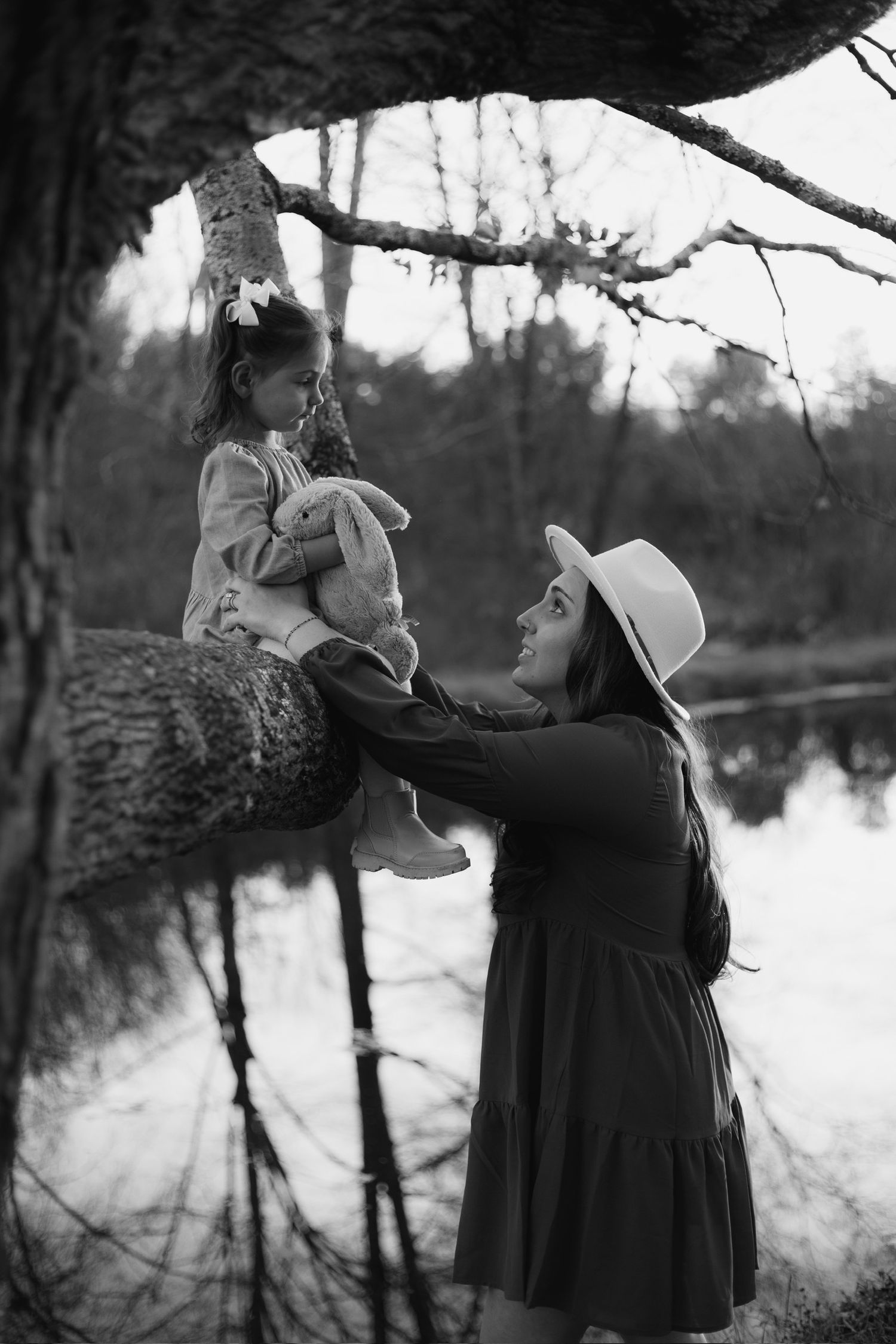 Black and white photo of mother in hat helping young girl sitting on tree branch holding stuffed animal near lake.
