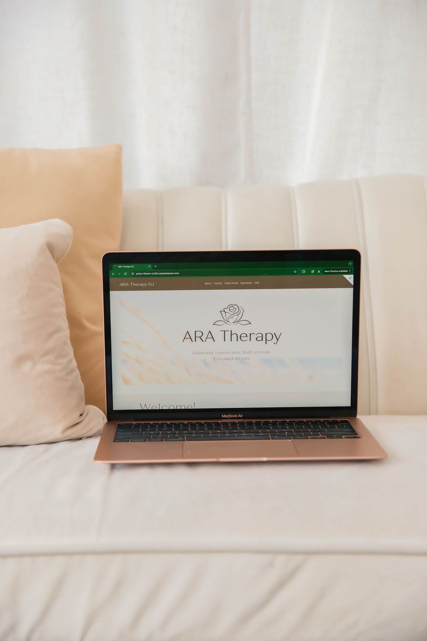 Rose gold MacBook laptop displaying ARA Therapy website on a cozy white sofa.