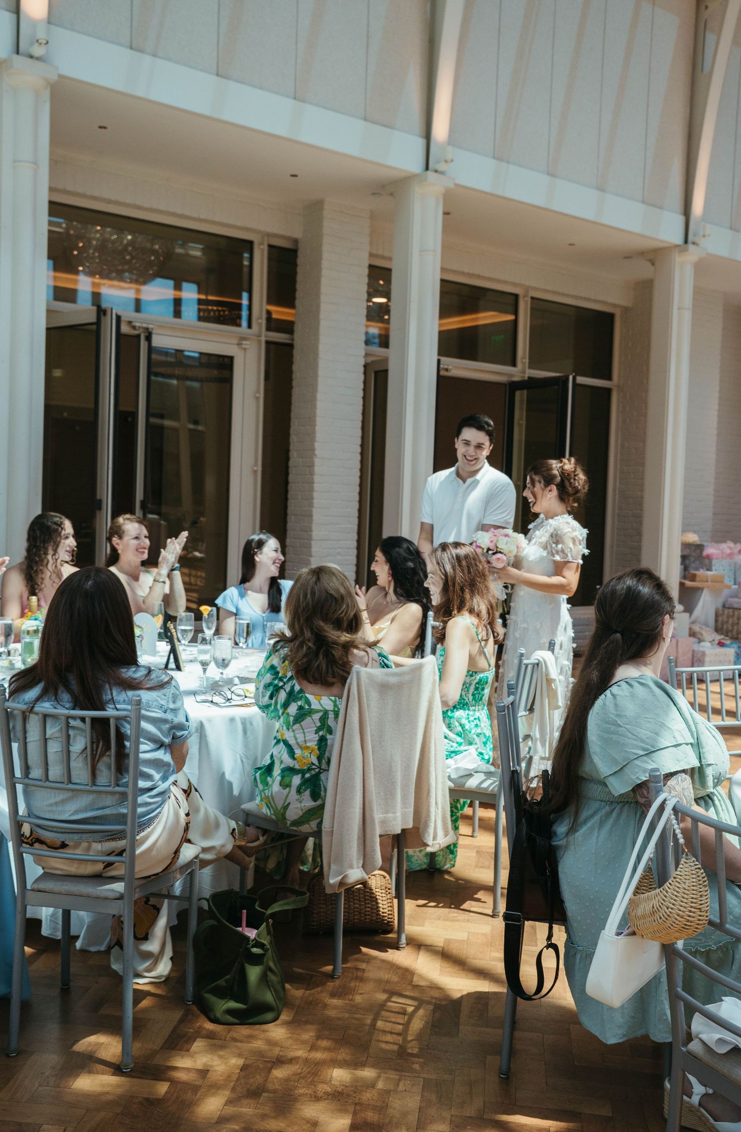 Guests gathered at elegant outdoor dining event, women in summer dresses seated at white-clothed tables in sunlit venue.
