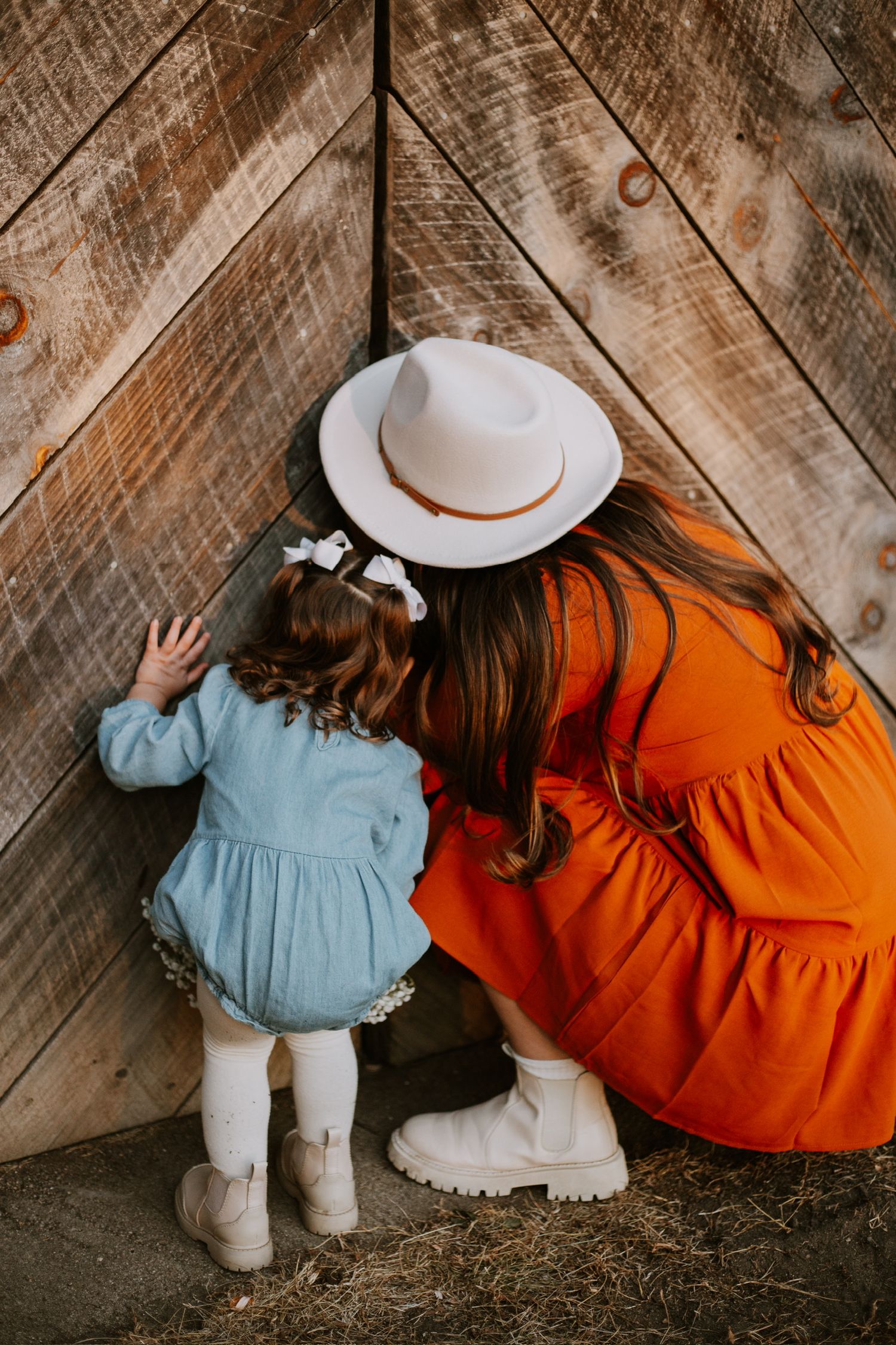 Woman in orange dress and white hat crouching with young girl in blue dress near rustic wooden barn wall.