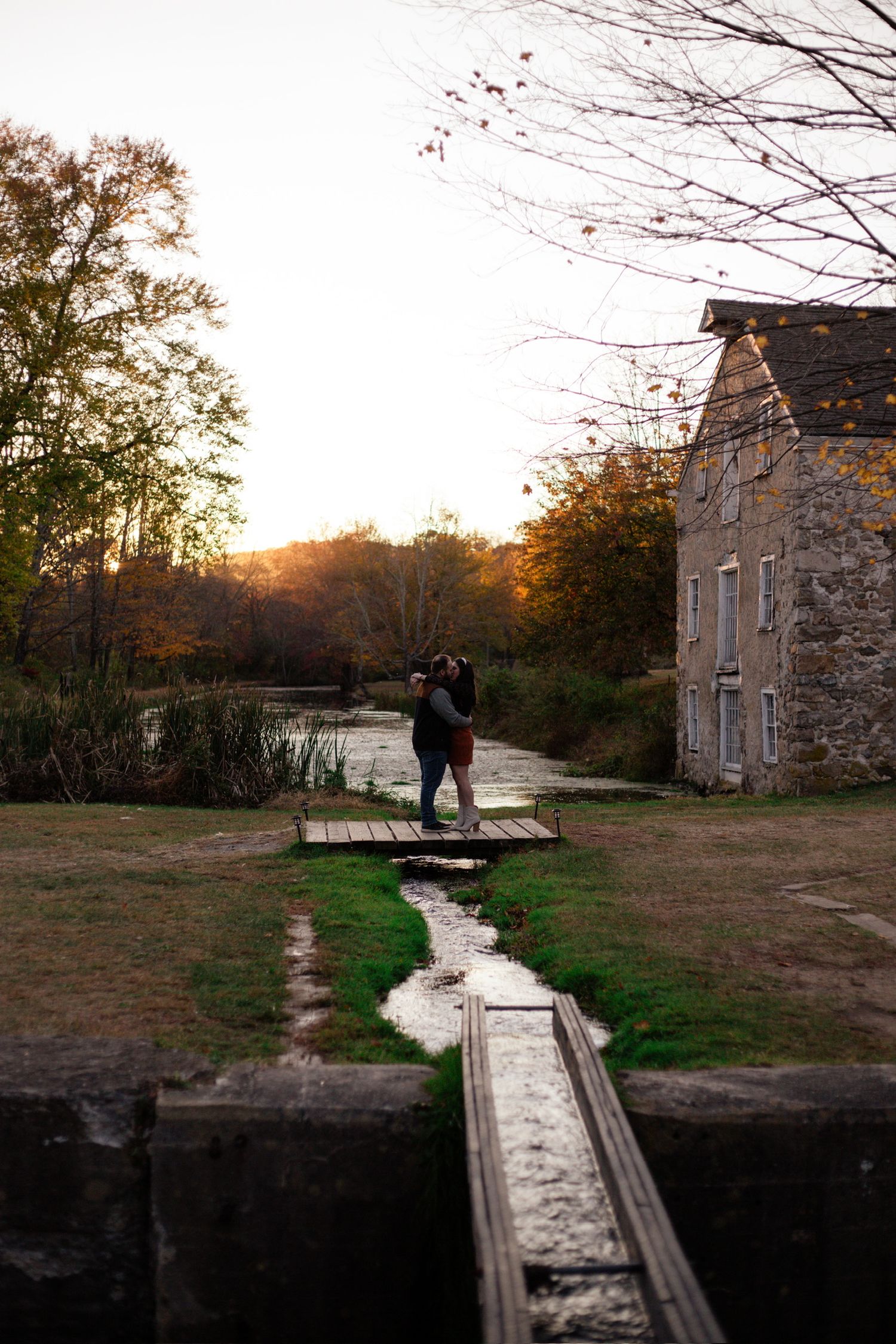 Couple embracing on a wooden bridge beside a historic stone mill at golden hour during autumn.