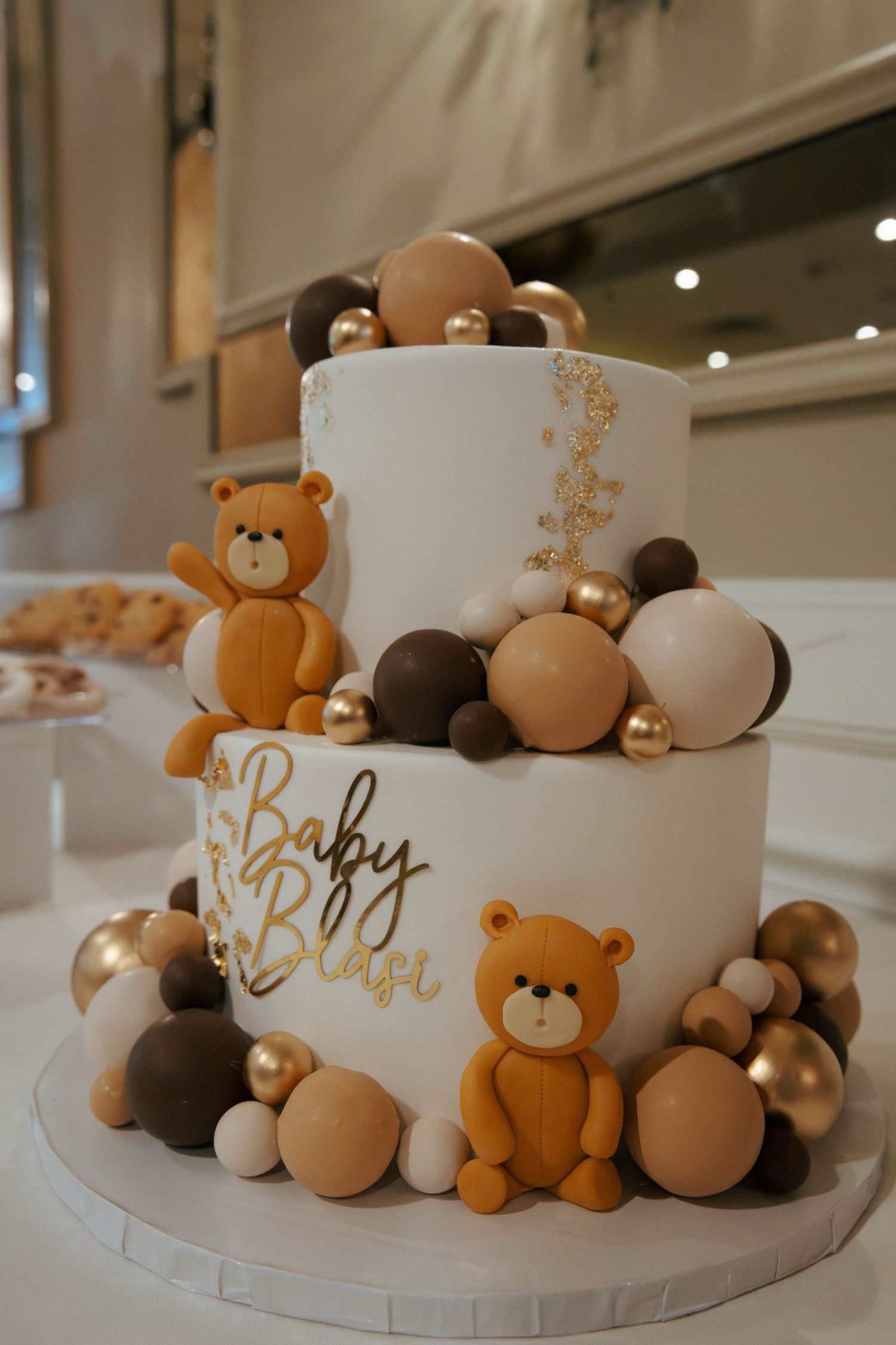 Two-tier white baby shower cake with teddy bear figurines, gold accents, and brown balloon decorations reading Baby Blasi.
