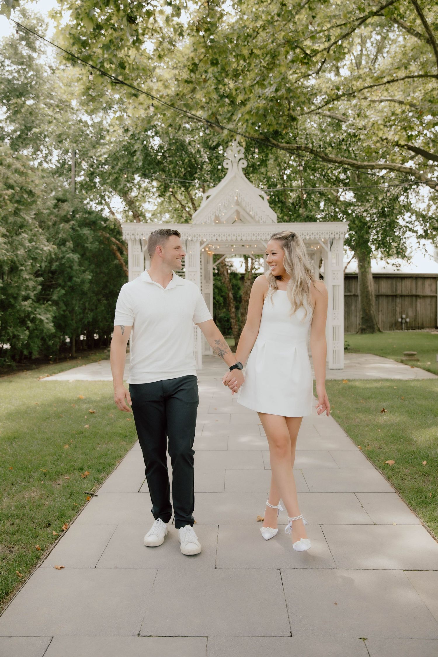 Couple in matching white outfits holding hands and walking along a stone path near a white gazebo.