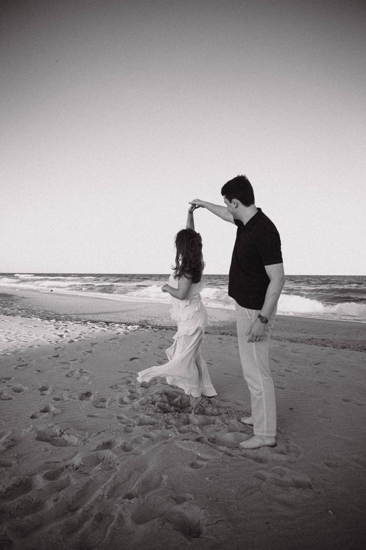 Black and white photo of a couple dancing on a beach, man spinning woman in white dress by the ocean.