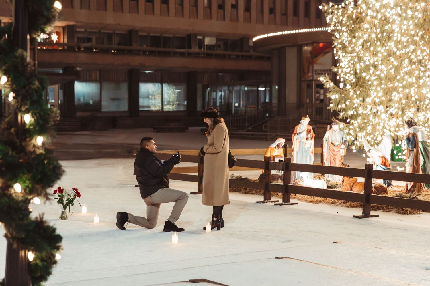 Nighttime ice rink proposal with man on one knee surrounded by festive holiday lights.