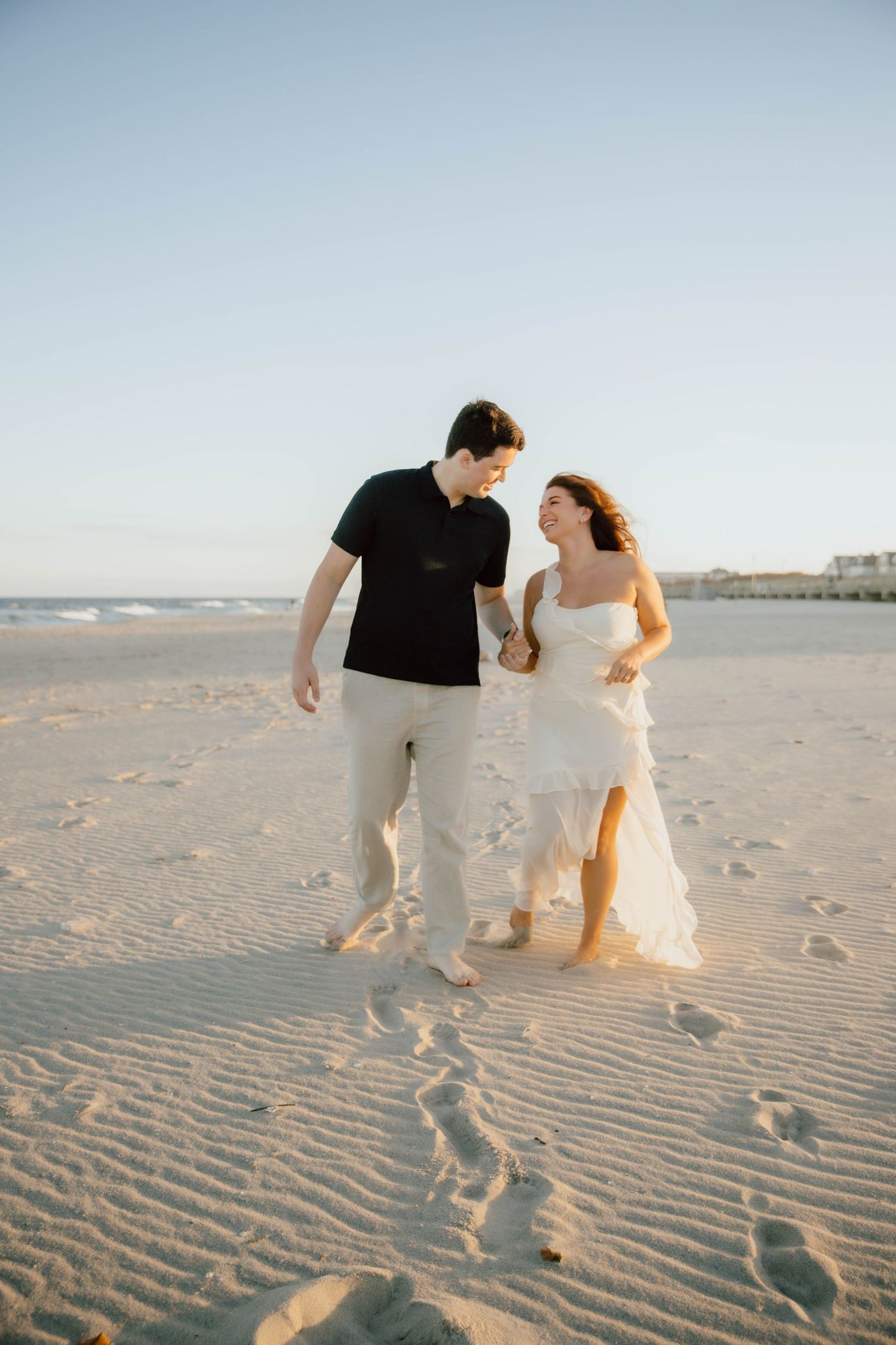 Couple walking hand-in-hand on a sandy beach at sunset, woman in flowing white dress.