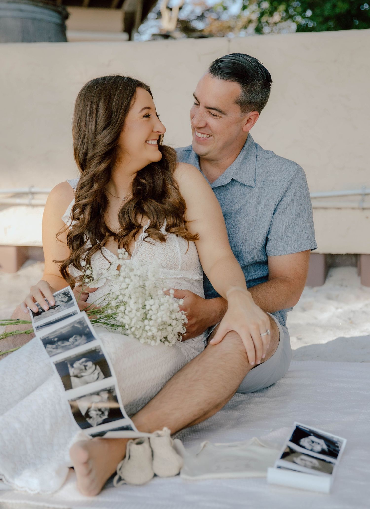 Happy couple sitting outdoors holding ultrasound photos and baby shoes during a maternity announcement.