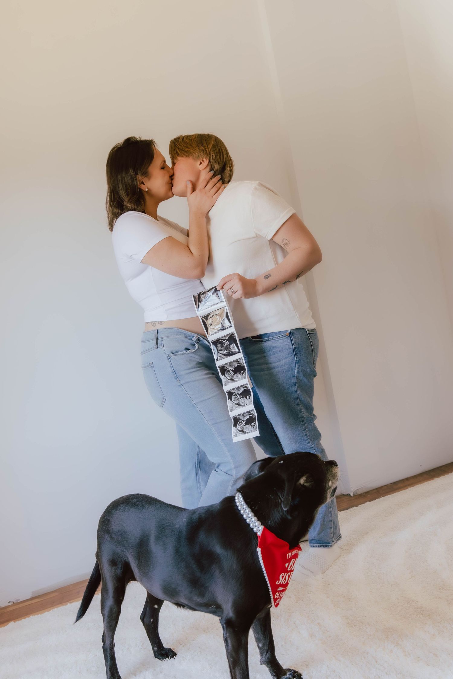 Couple kissing while holding ultrasound photos, with a black dog wearing a red bandana in a pregnancy announcement.
