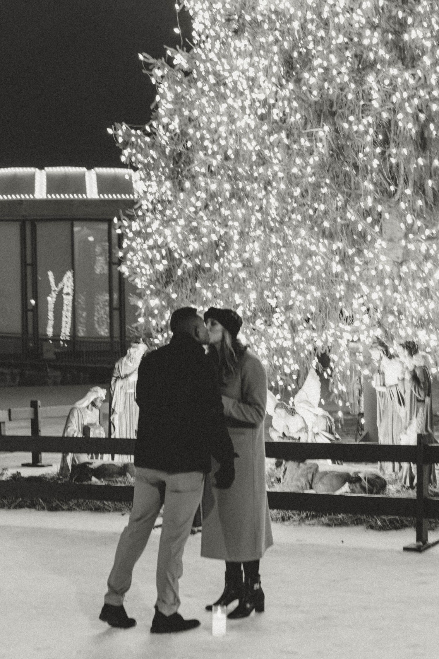 Couple kissing in front of a large illuminated Christmas tree at night in a black and white photo.