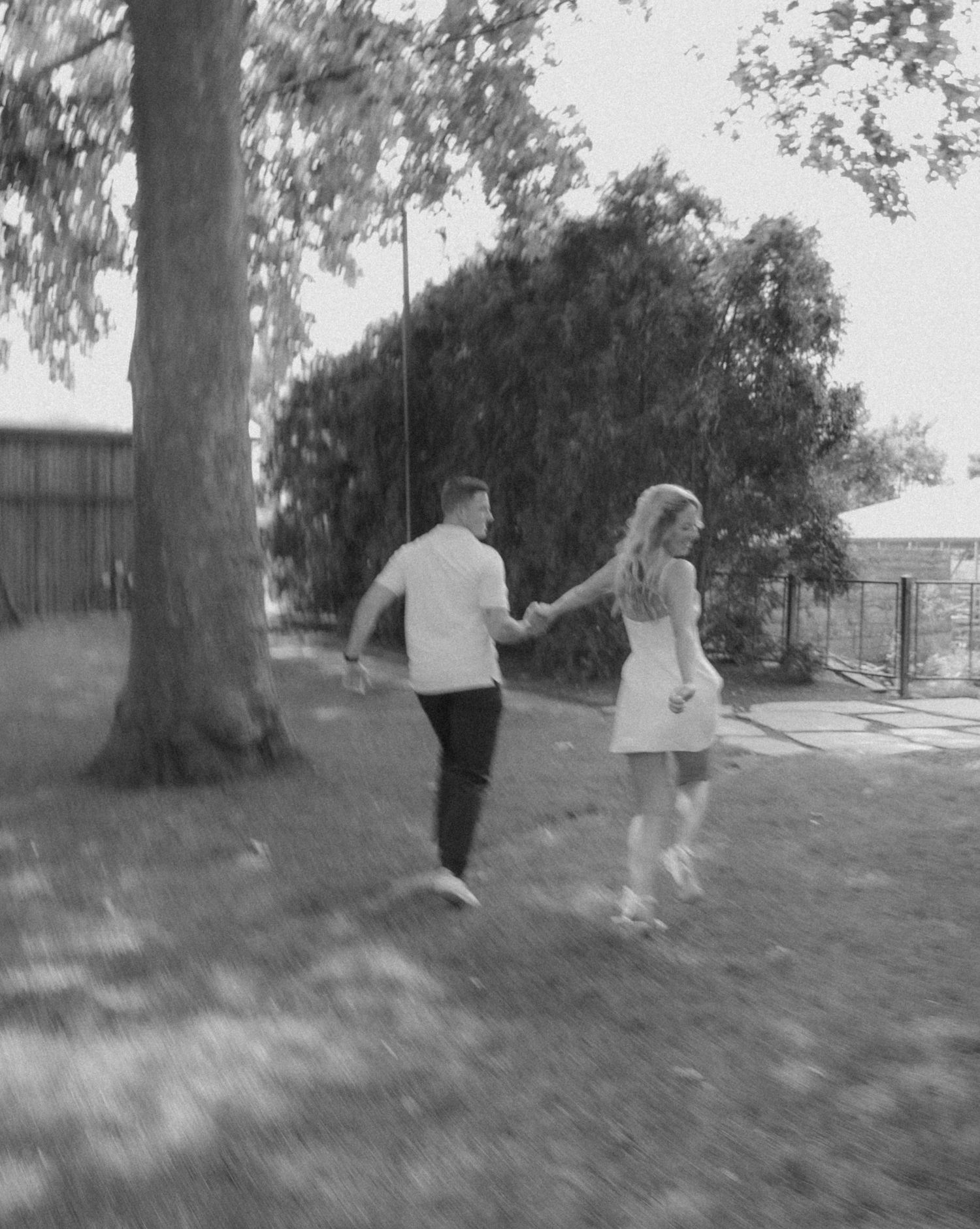 Black and white photo of a joyful couple running hand in hand through a sunny backyard.