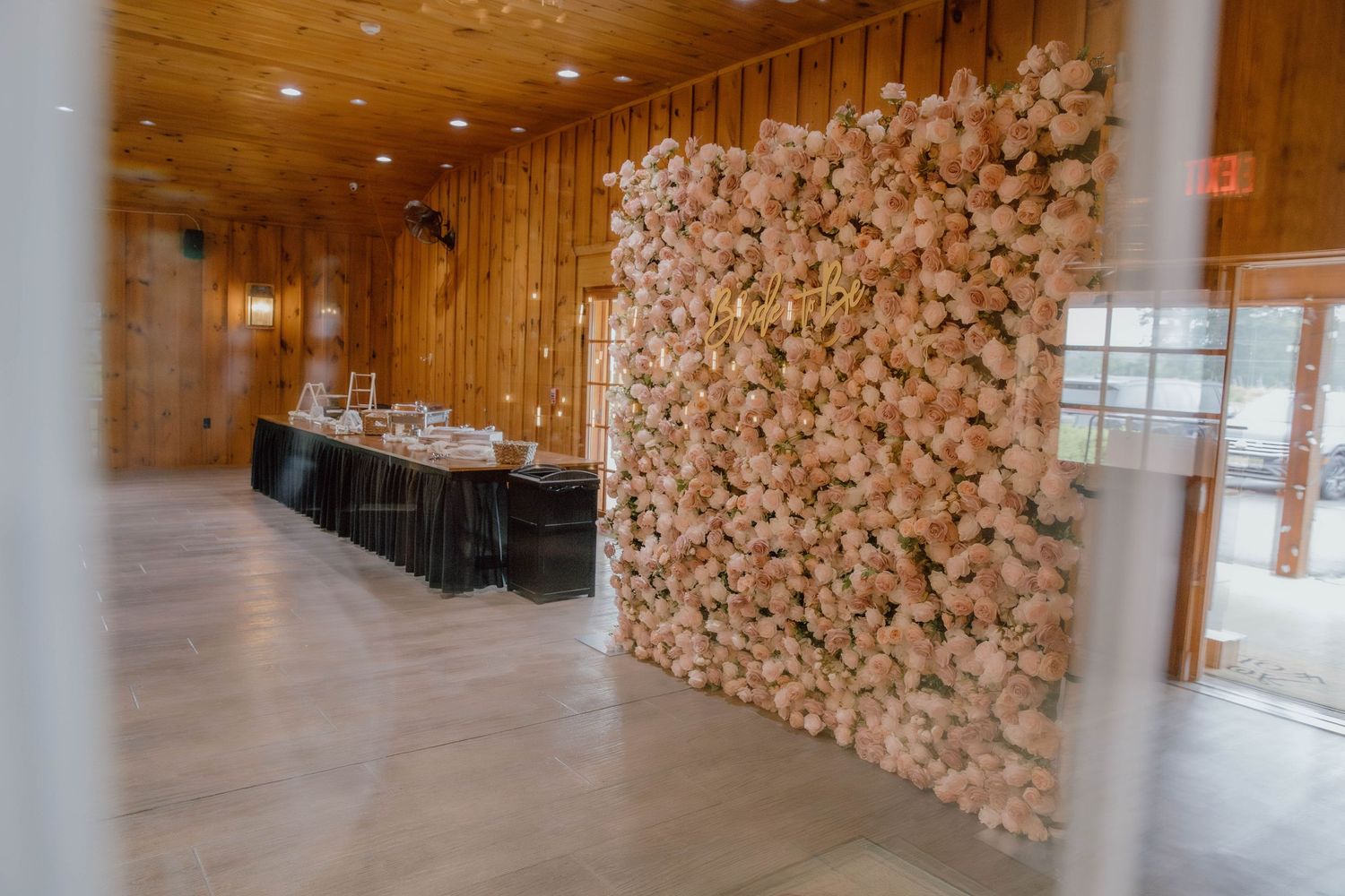 Elegant floral wall backdrop beside draped banquet table in rustic wood-paneled event venue.