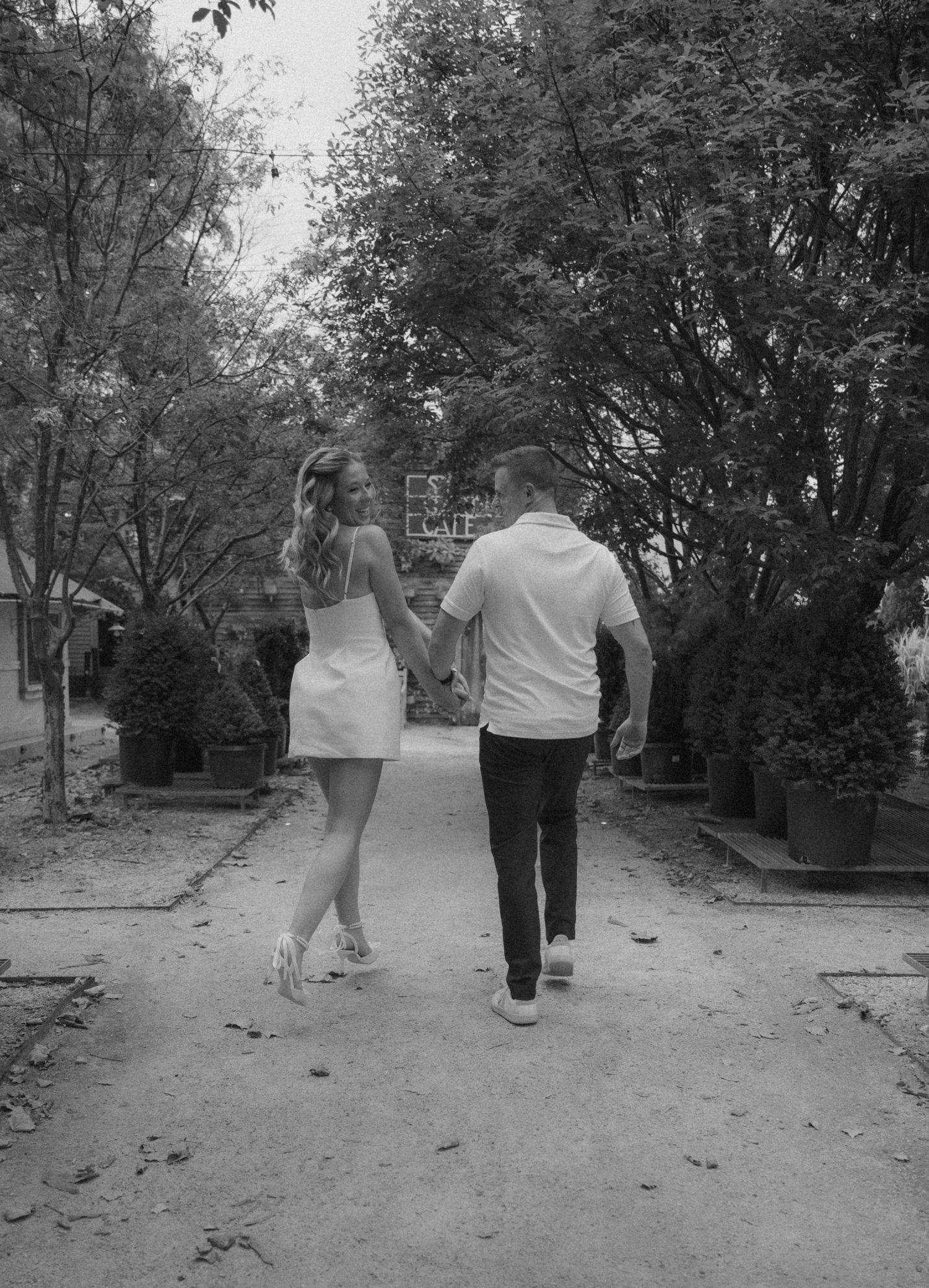 Black and white photo of a couple holding hands walking down a tree-lined path, woman in white mini dress.