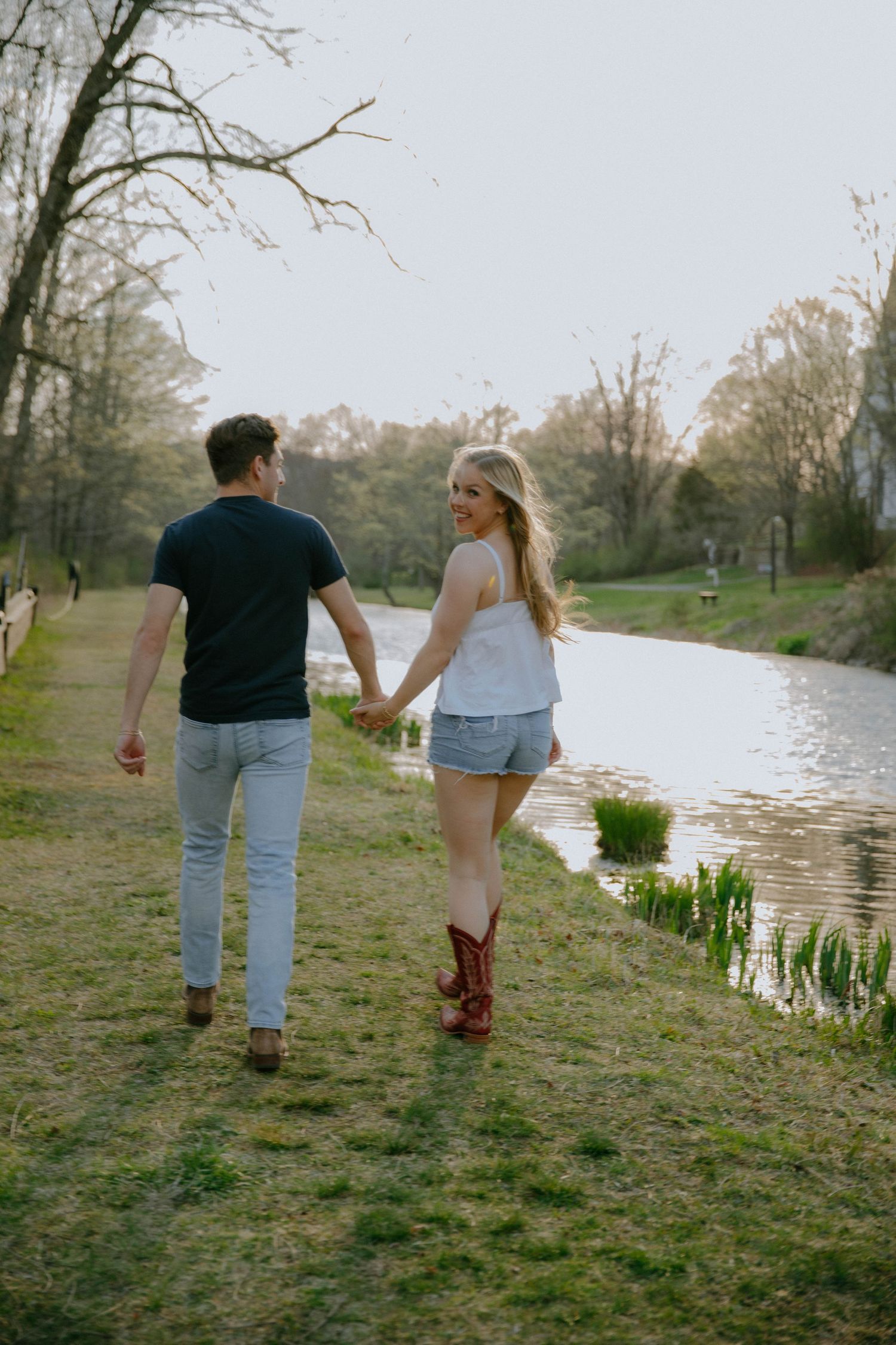 Couple holding hands walking along a scenic pond path, woman in cowboy boots and denim shorts looking back smiling.