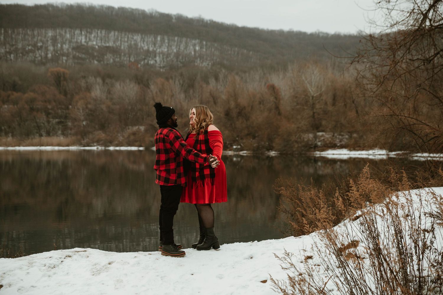 Couple in matching red plaid outfits standing by a snowy lakeside in winter landscape.