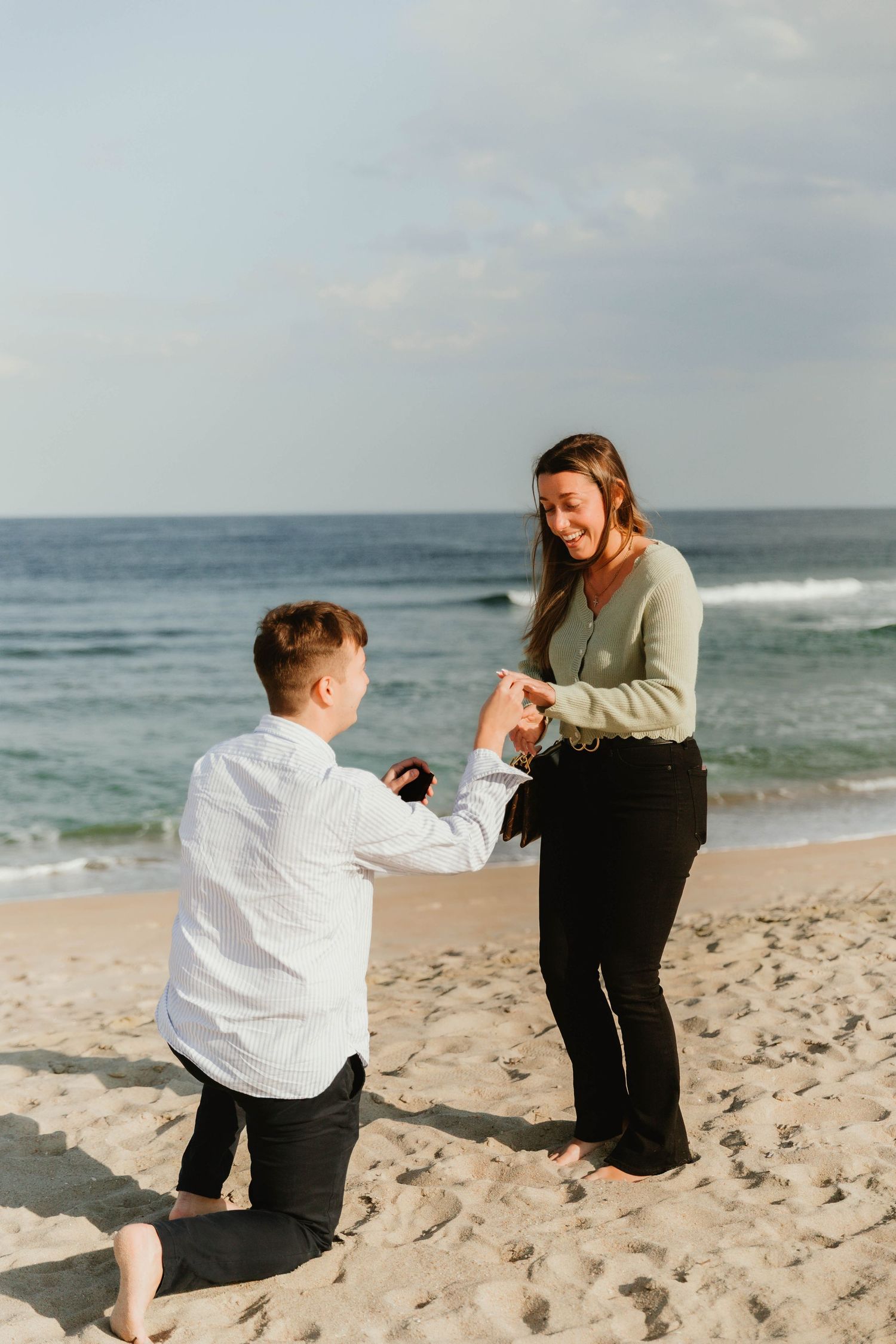 Man proposing on one knee to smiling woman on a sandy beach with ocean waves behind them.