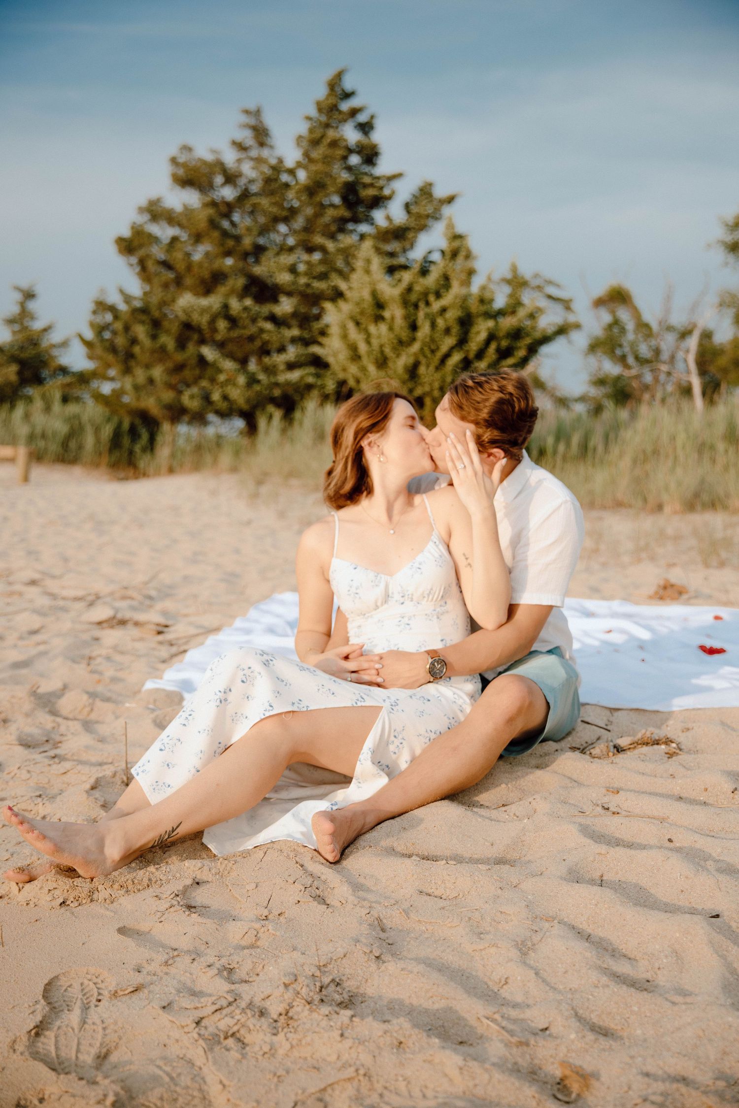 Romantic couple sharing a kiss while sitting on a sandy beach blanket with evergreen trees in the background.