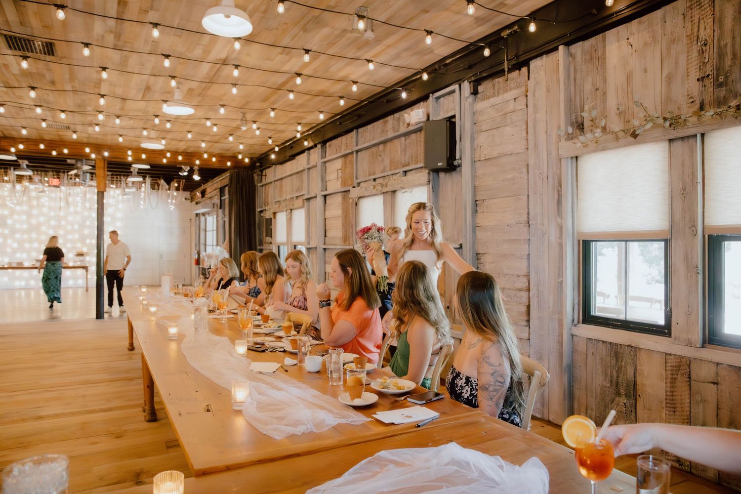 Women gathered at a long wooden table in a rustic barn venue with string lights for a bridal shower luncheon.