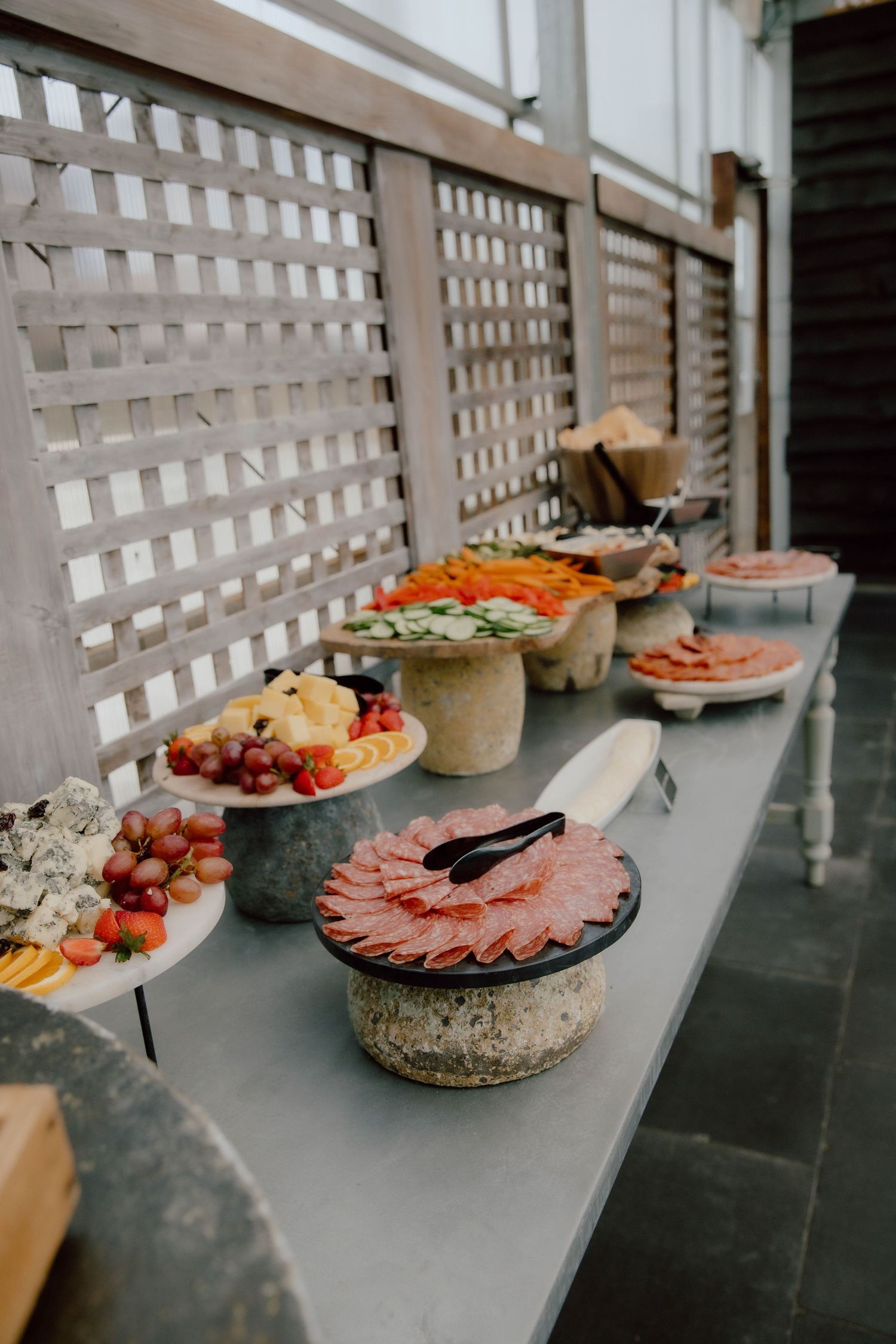 Elegant outdoor buffet table with charcuterie, fresh fruits, and cheese displayed on stone pedestals.