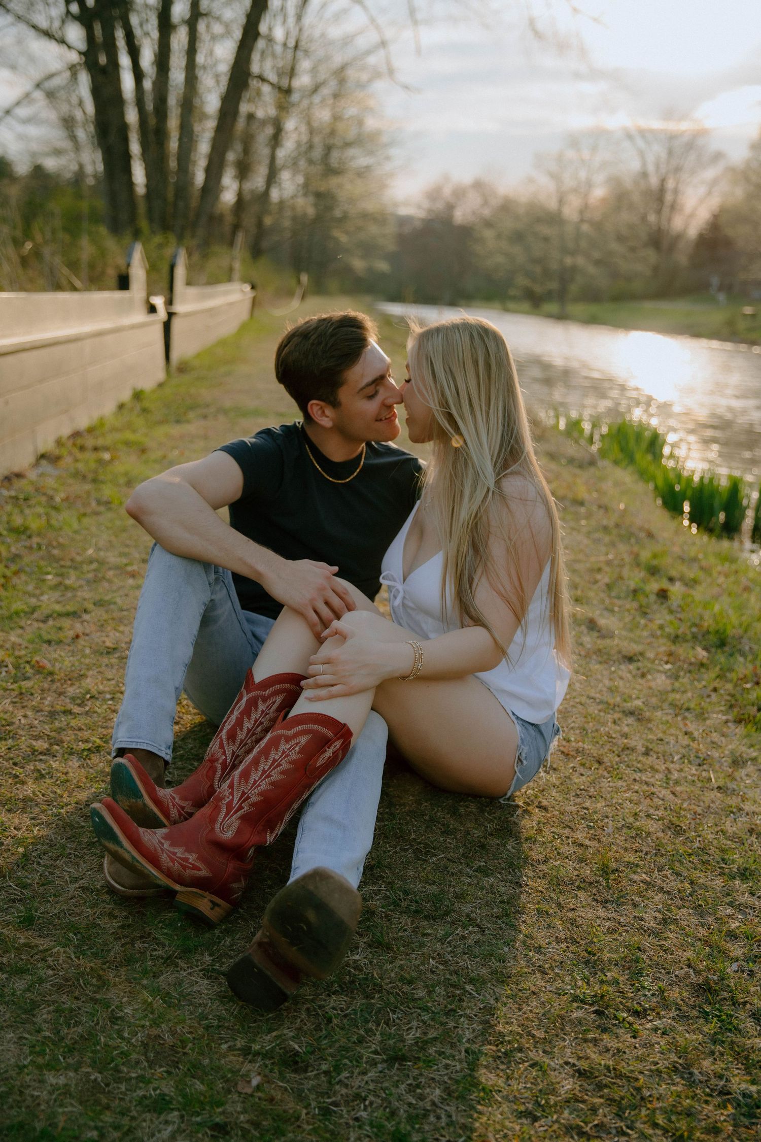 Young couple about to kiss while sitting on grass beside a peaceful pond at golden hour sunset.