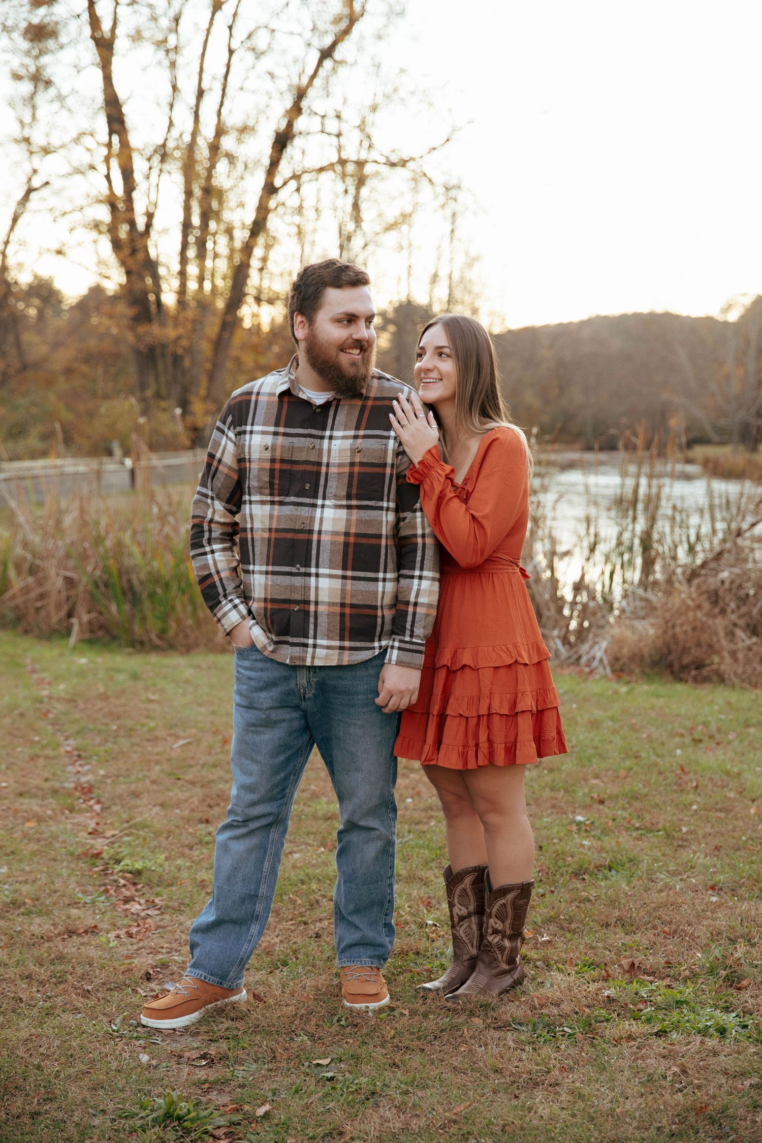 Couple holding hands outdoors in fall, man in plaid shirt and jeans, woman in rust orange dress and cowboy boots.