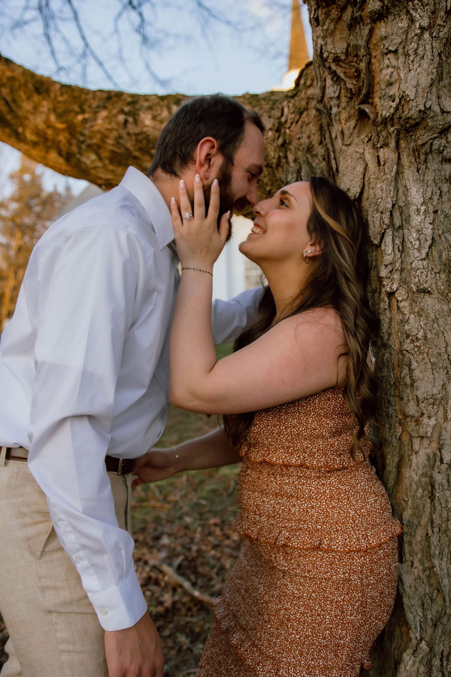 Romantic couple almost kissing while leaning against large tree trunk in golden autumn light.