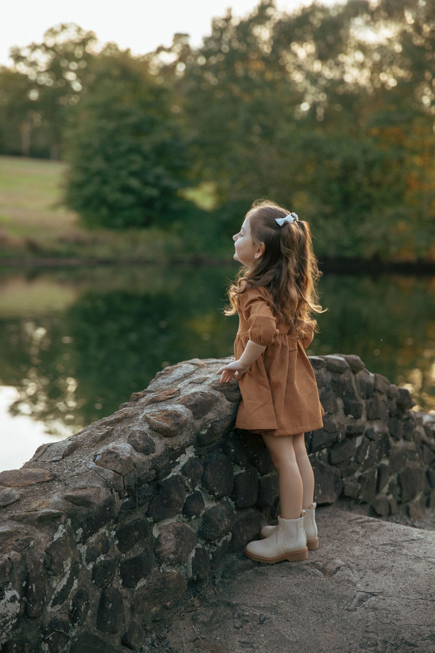 A little girl in a brown dress and white boots stands by a stone wall overlooking a serene pond in a lush park.