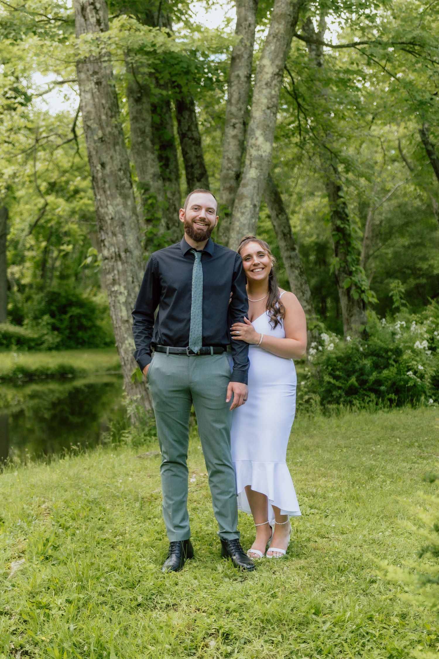 A smiling couple poses outdoors on green grass, man in black shirt and tie, woman in white dress, with trees behind.