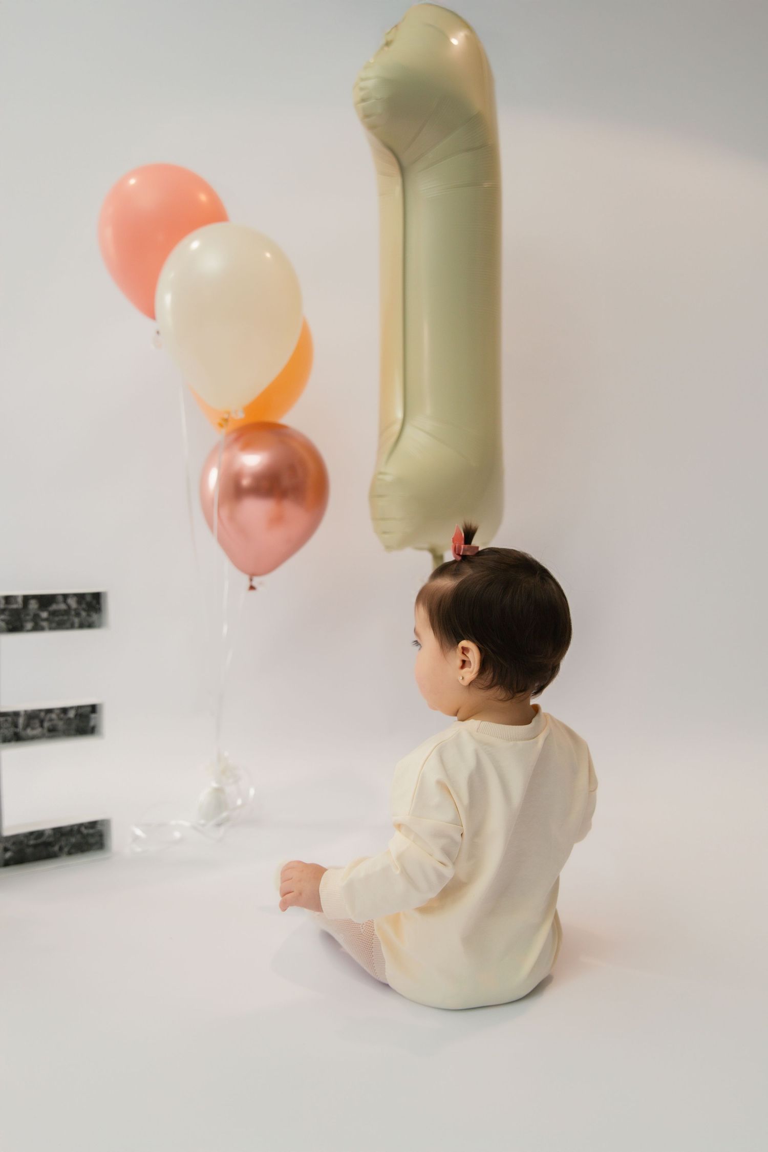 Baby in white onesie sitting on floor with colorful balloons and number 1 balloon for first birthday celebration.