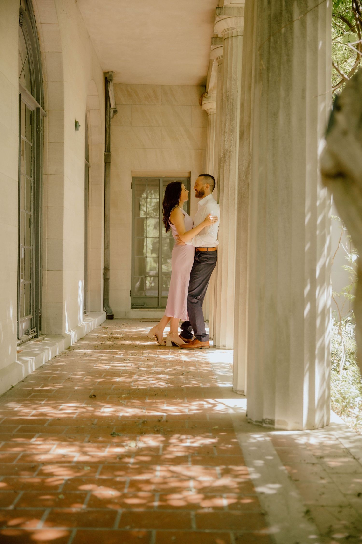 Couple embracing on a sunlit brick colonnade with white columns and dappled light.