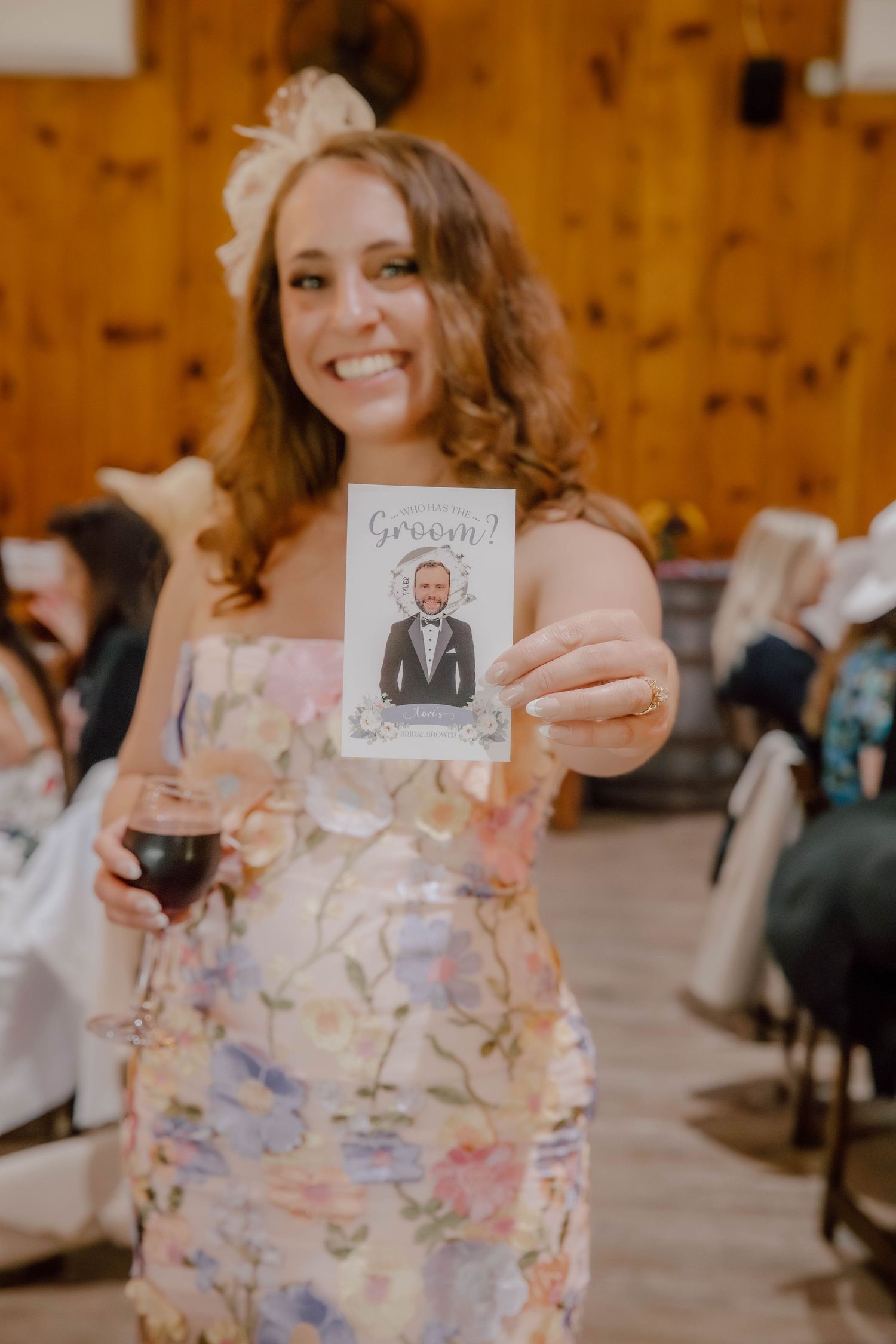 Smiling woman in floral dress holding groom caricature card and wine glass at wedding reception.