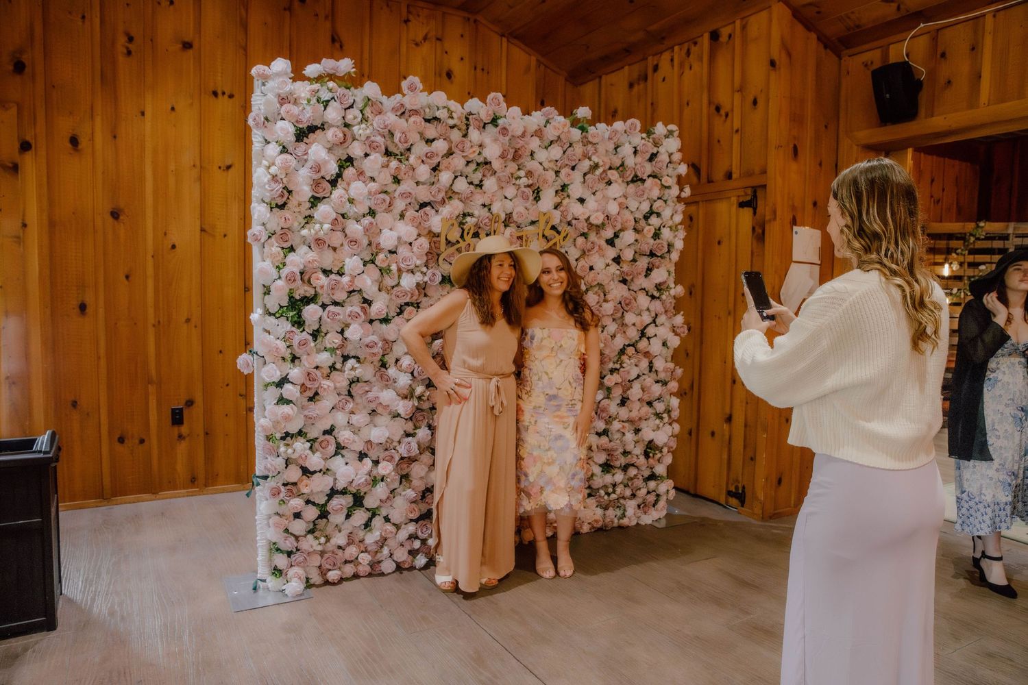 Two women pose in front of a pink floral wall backdrop while a third photographs them at an elegant event.