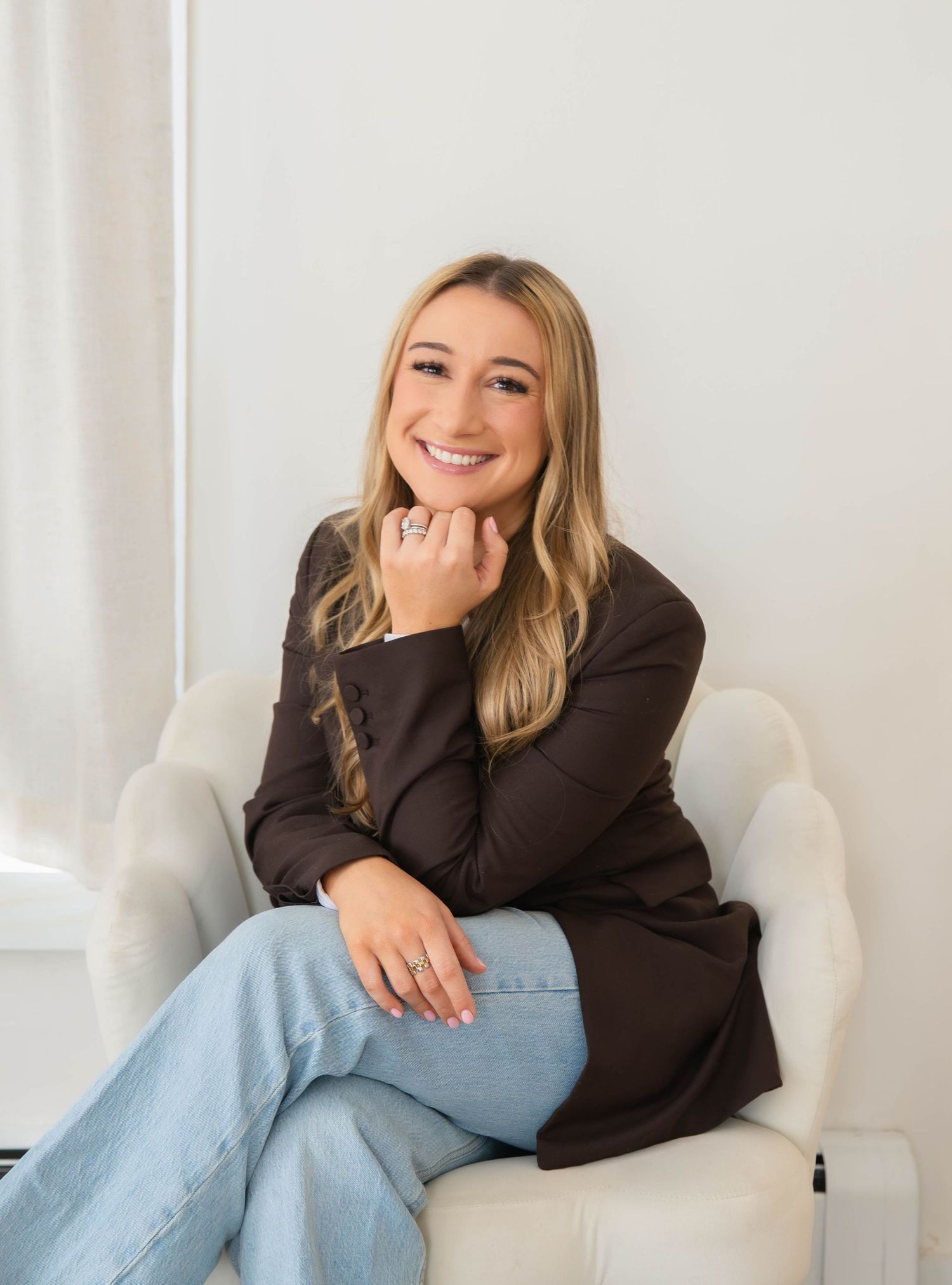 Smiling blonde woman in brown blazer and jeans seated in white armchair indoors.