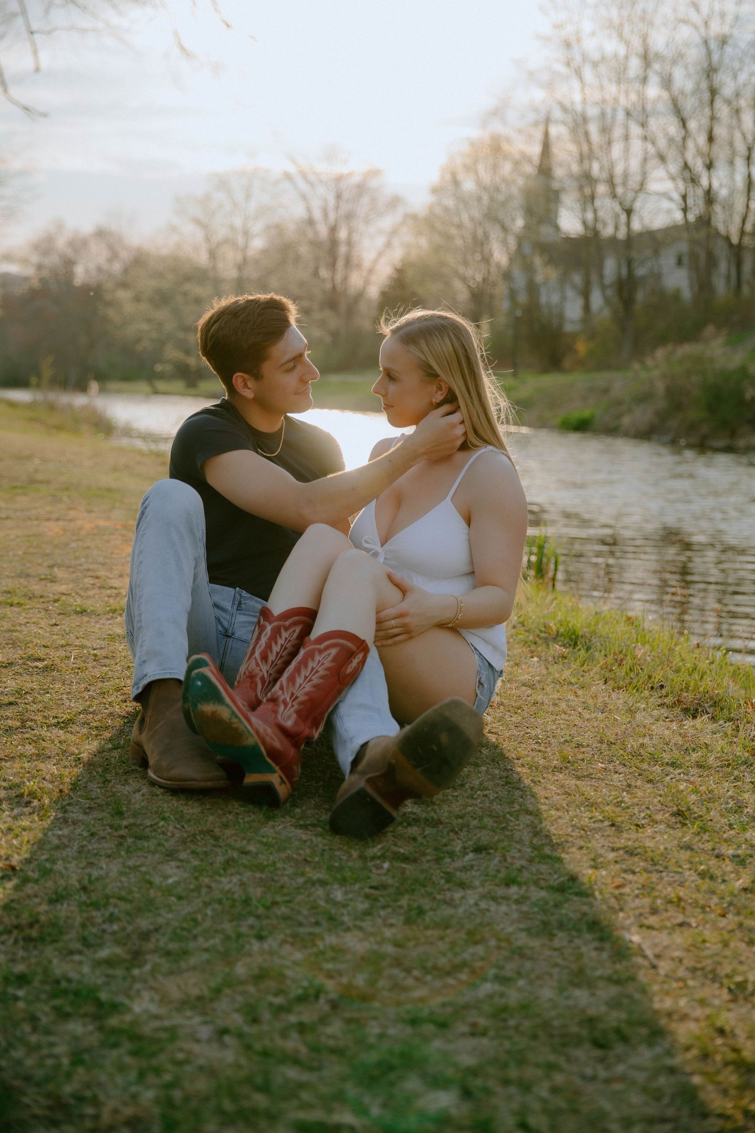 Couple sitting together on grass by a river at golden hour, gazing into each other's eyes.