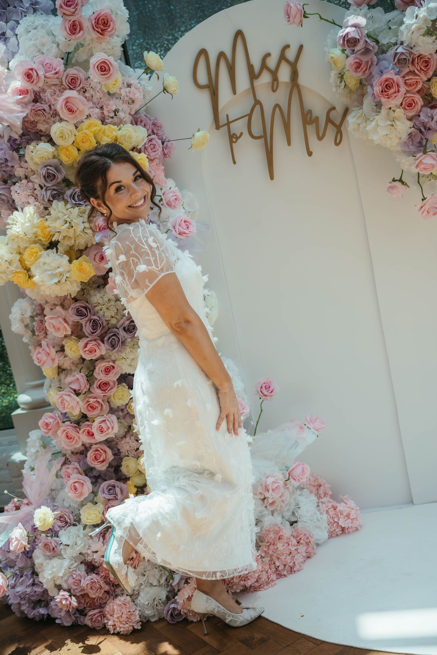 Bride in white lace dress posing by floral arch with Miss to Mrs sign at bridal shower.