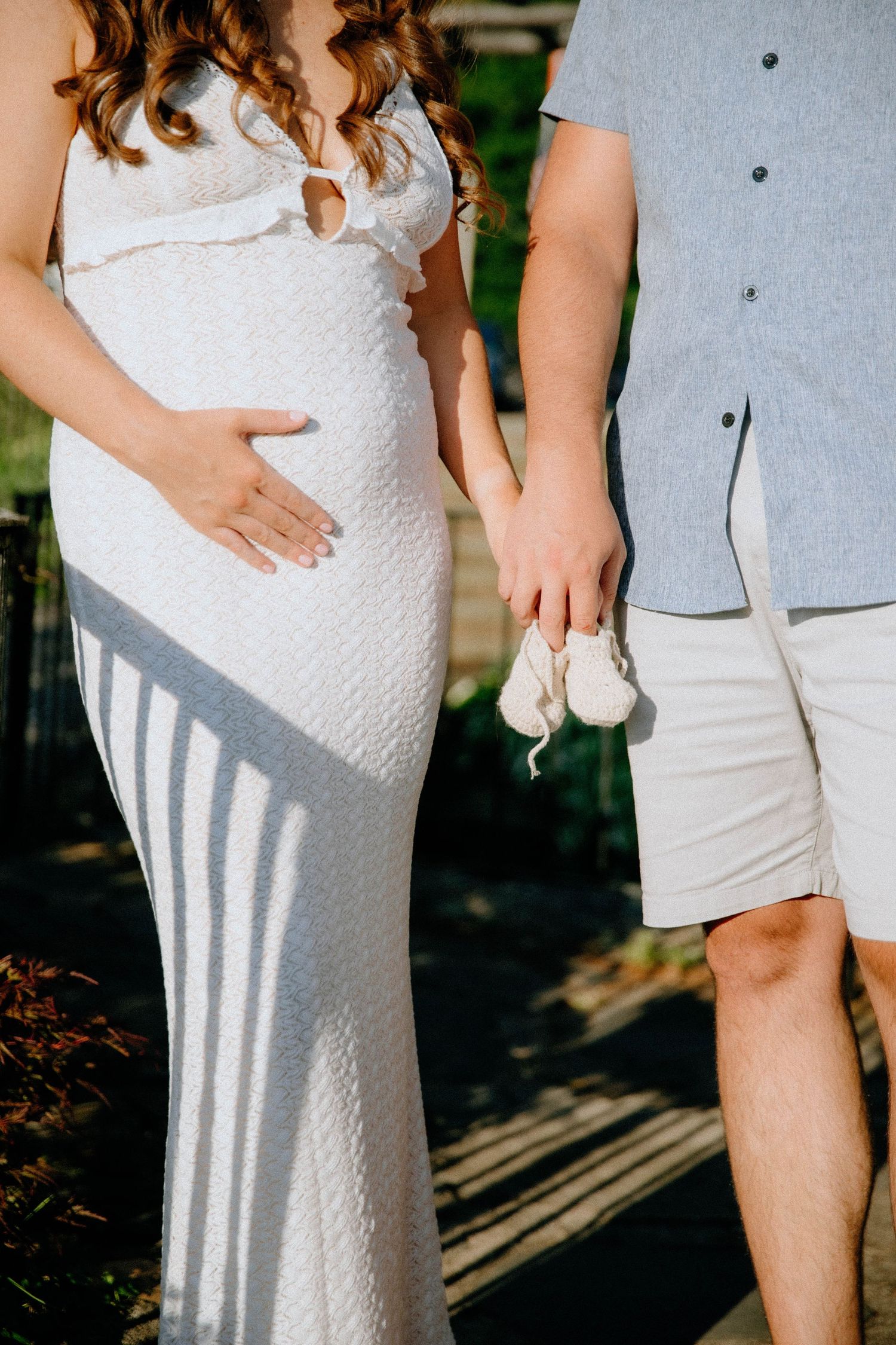 Pregnant woman in white dress holding tiny baby shoes with partner in sunlit outdoor setting.